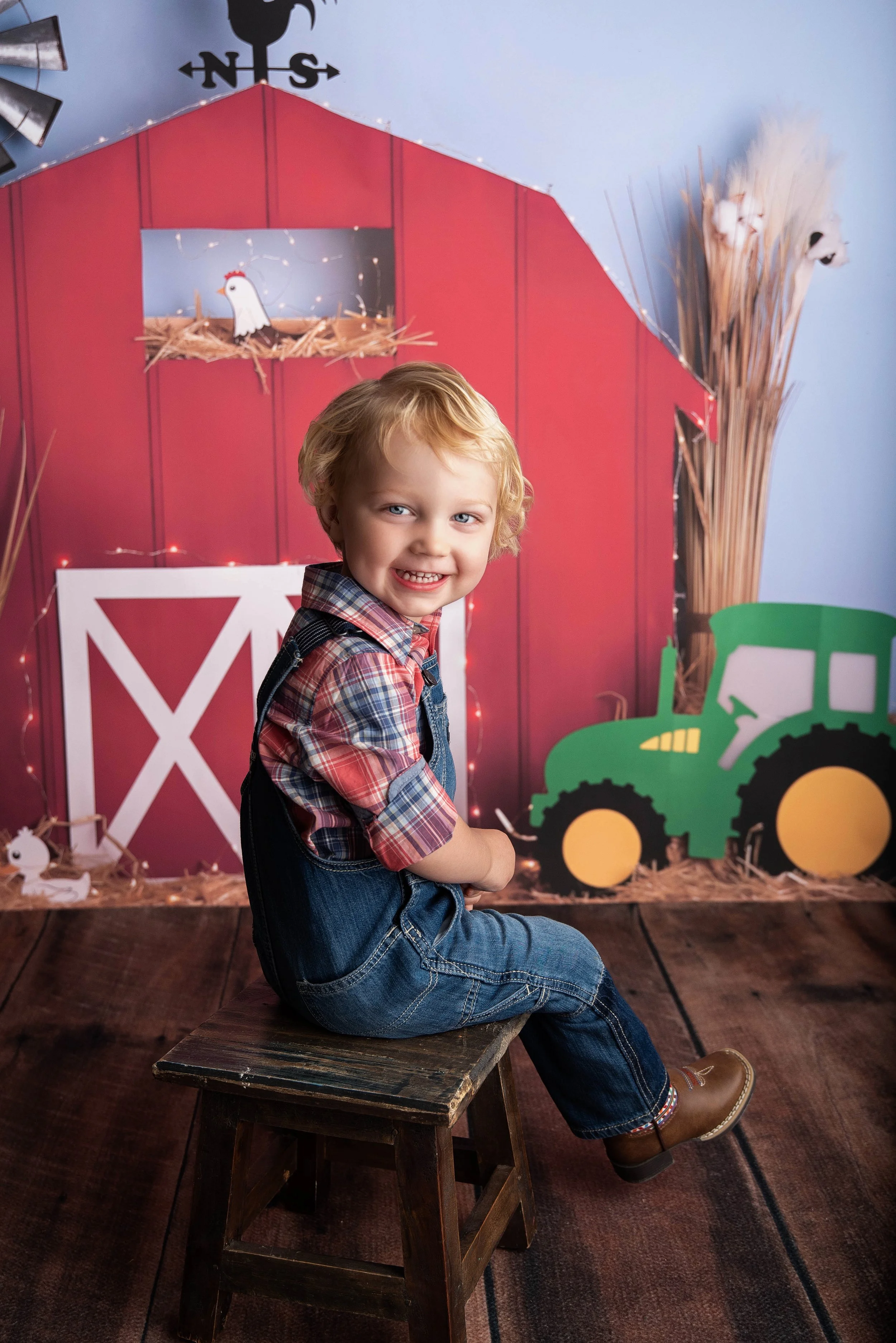 A young boy with blonde hair, wearing a plaid shirt, jeans, and brown boots, smiling and sitting on a small wooden stool in front of a farm-themed backdrop with a red barn, a green tractor, and farm animals.
