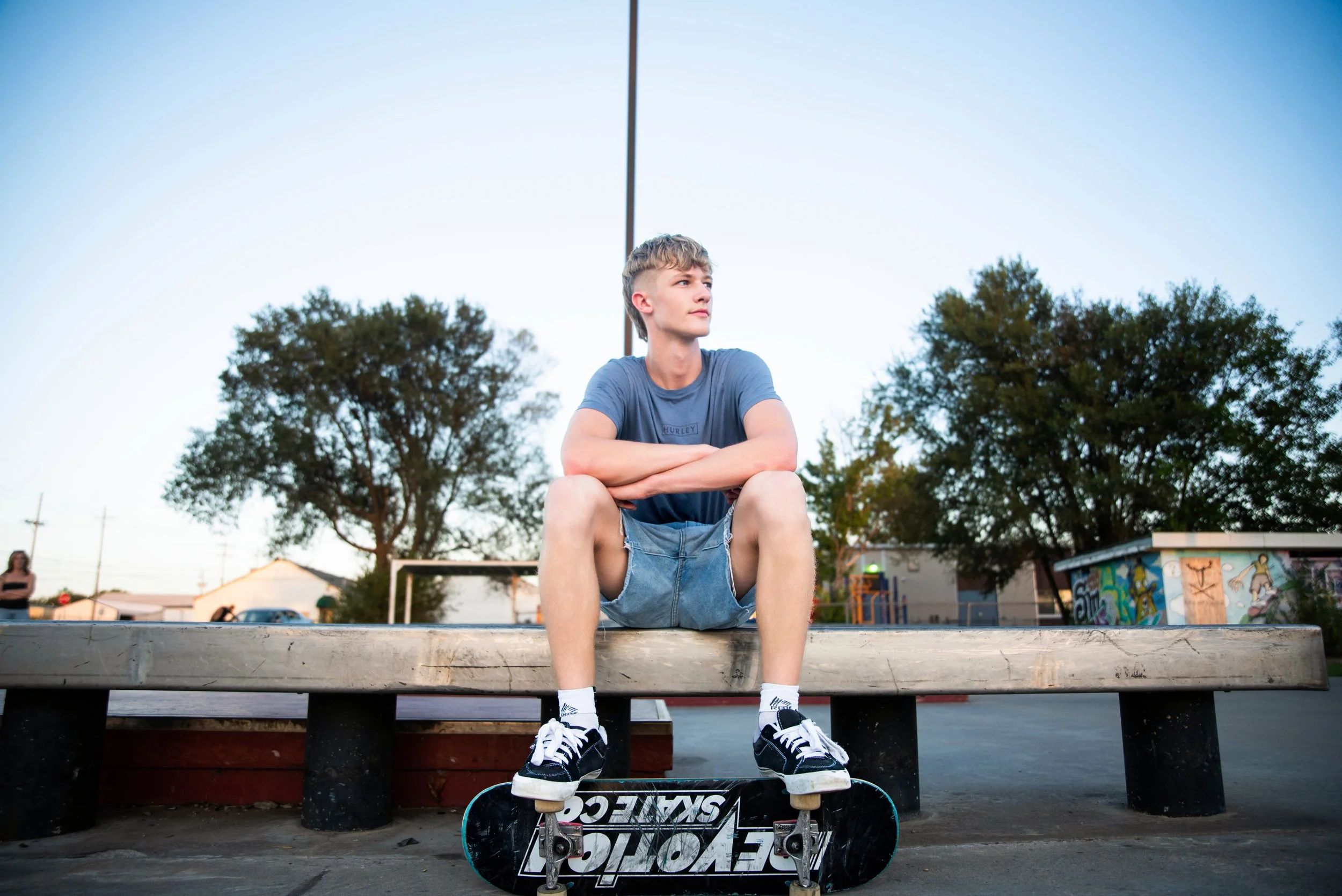 Young man sitting on a park bench with skateboards at his feet, arms crossed, looking to the side, with trees and buildings in the background during evening.