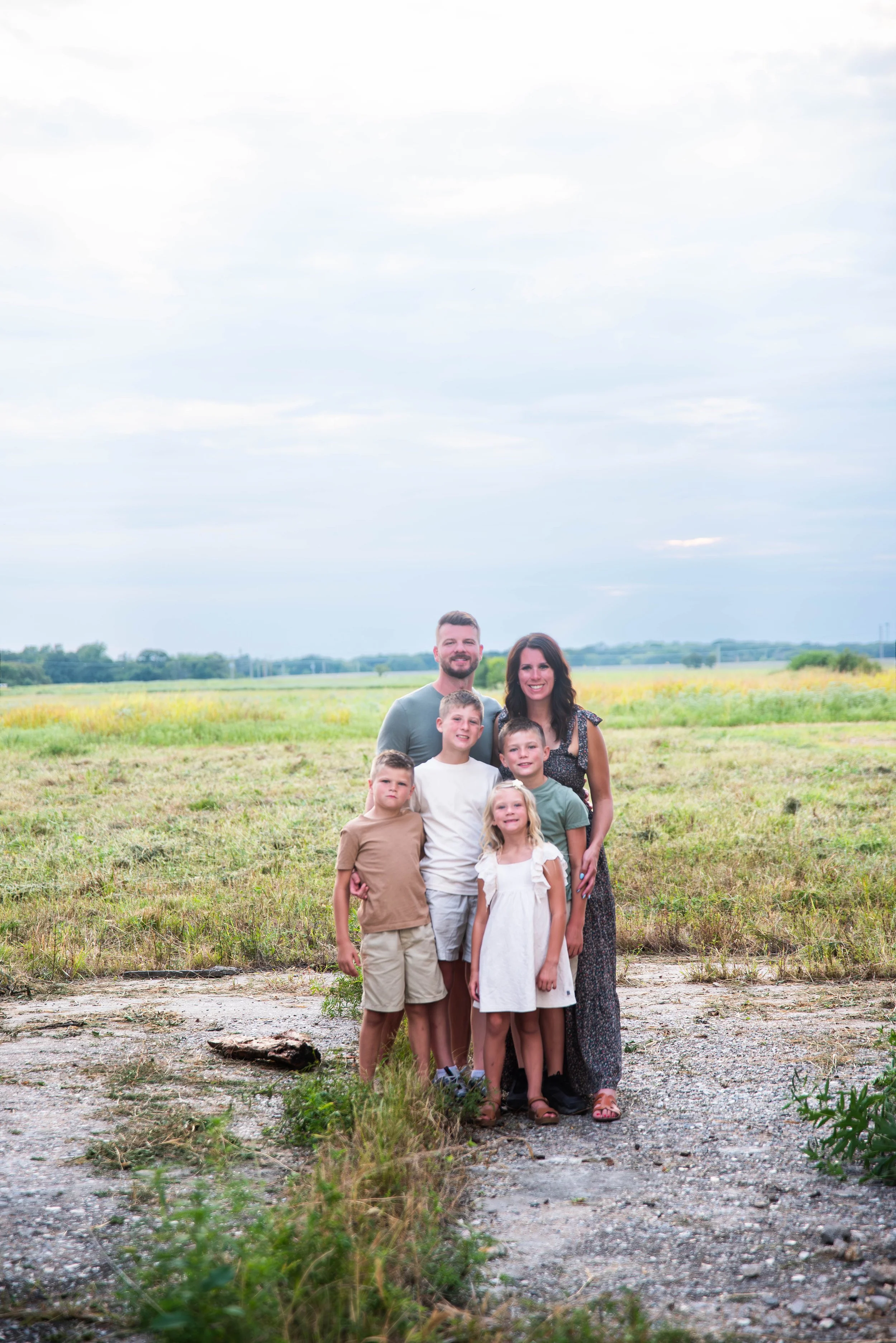 A family of six standing outdoors in a grassy field with a cloudy sky overhead.