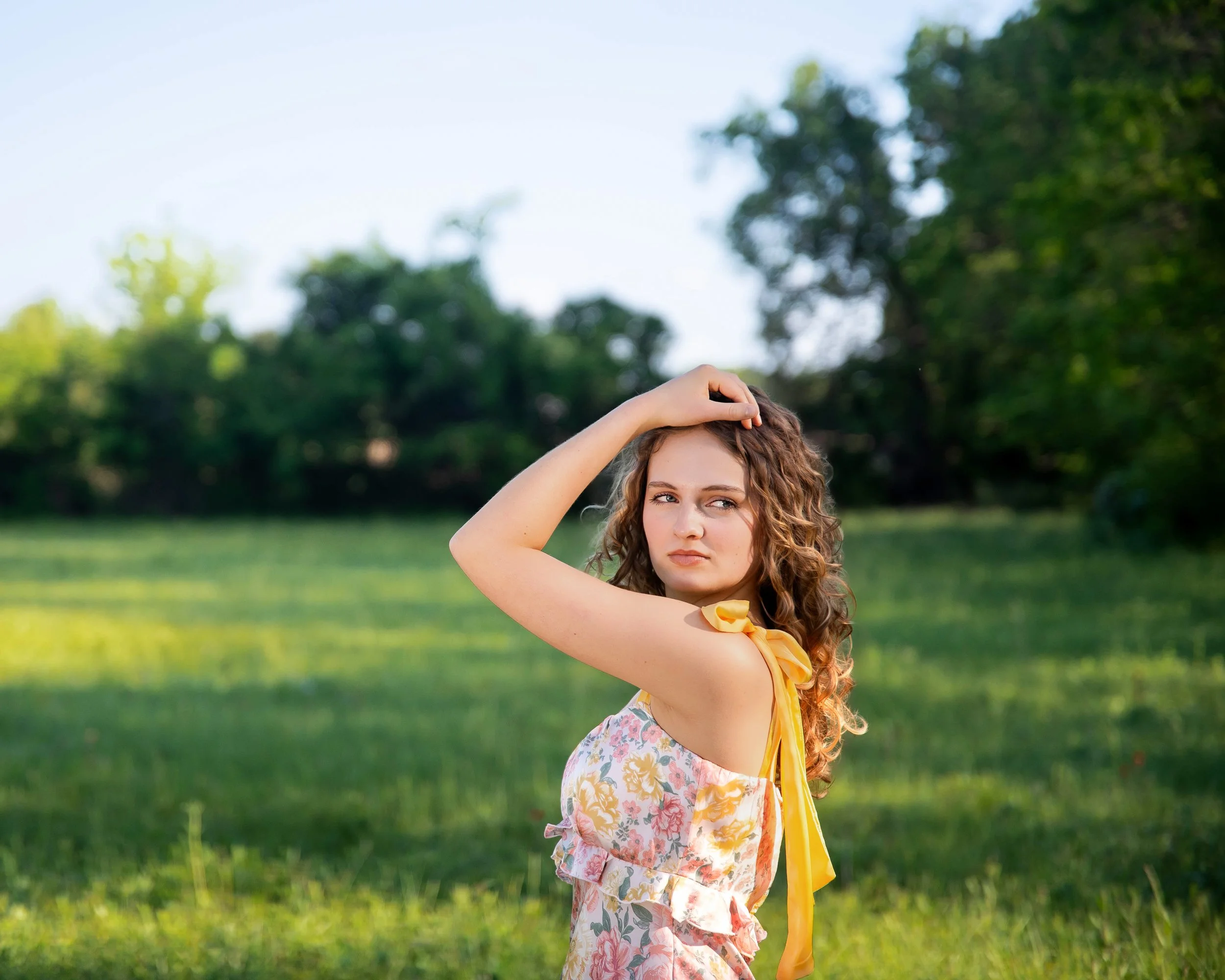 A young woman with curly hair standing outdoors in a grassy field, looking to the side with a serious expression, wearing a floral dress with yellow ribbon details.