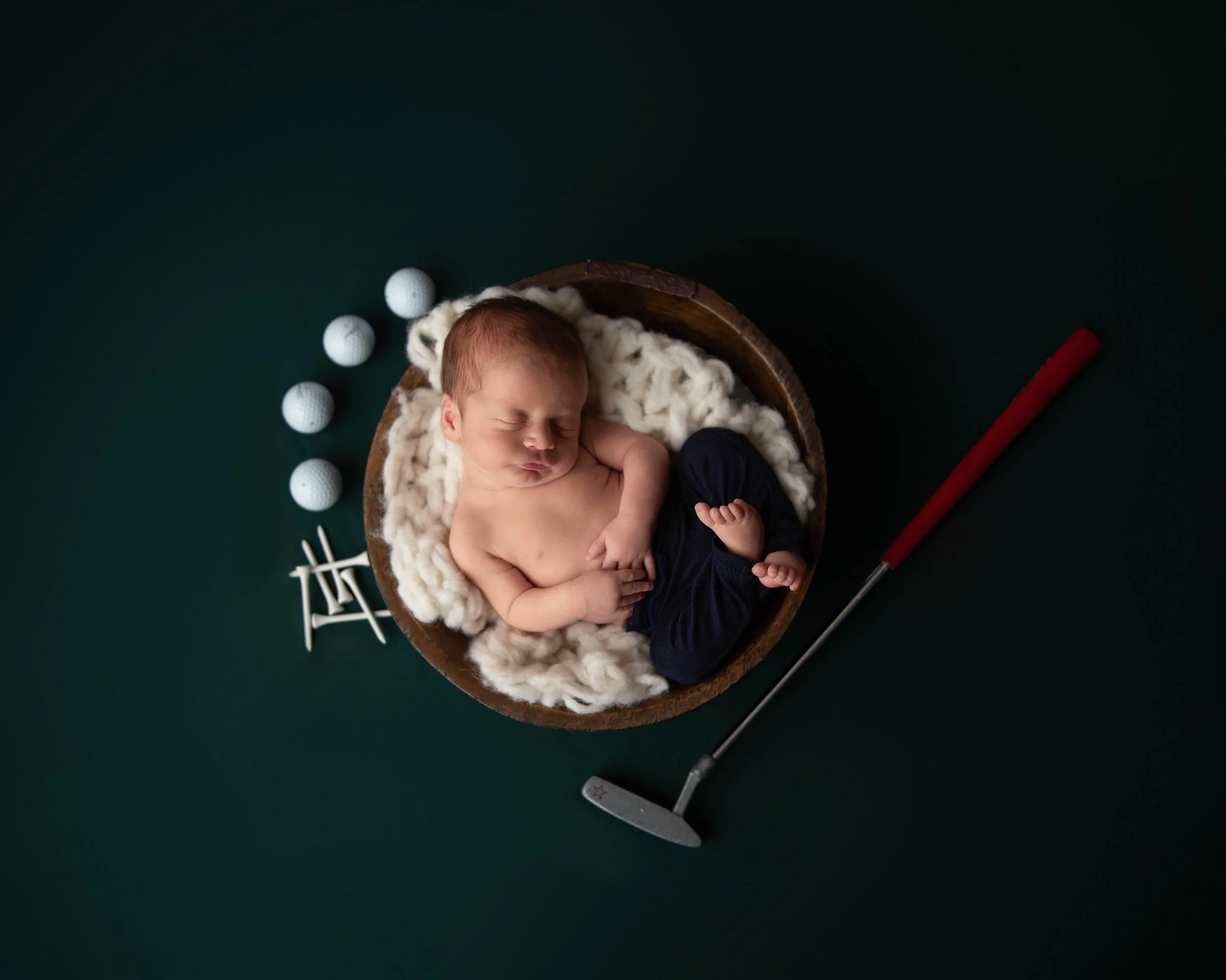 A sleeping baby lying in a circular wooden bowl on a dark green surface, wrapped in a soft white blanket, with golf balls and a mini golf putter nearby.
