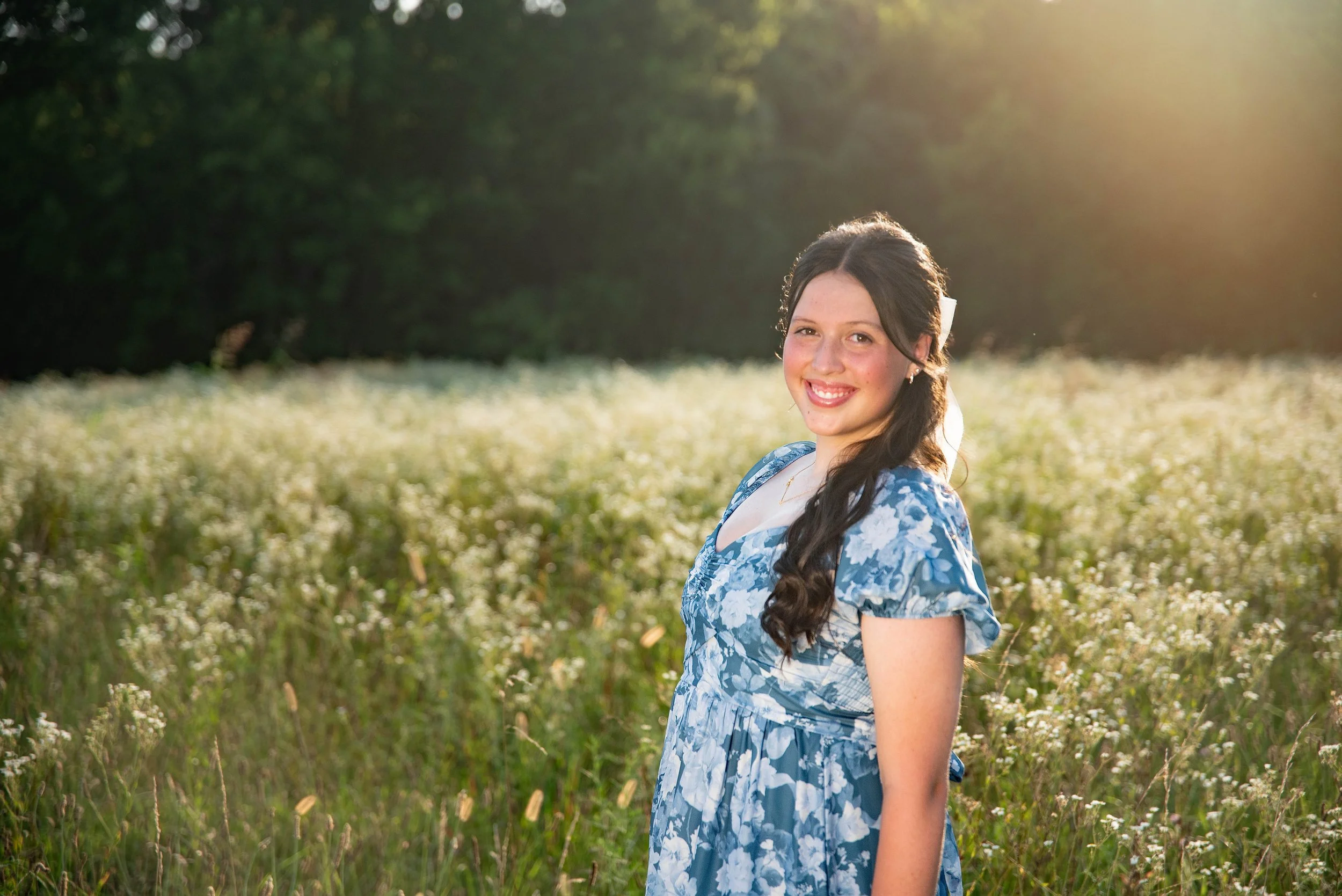 A young woman with dark brown hair, wavy and tied in a low ponytail, standing in a field of white wildflowers during sunset, wearing a blue floral dress and smiling.