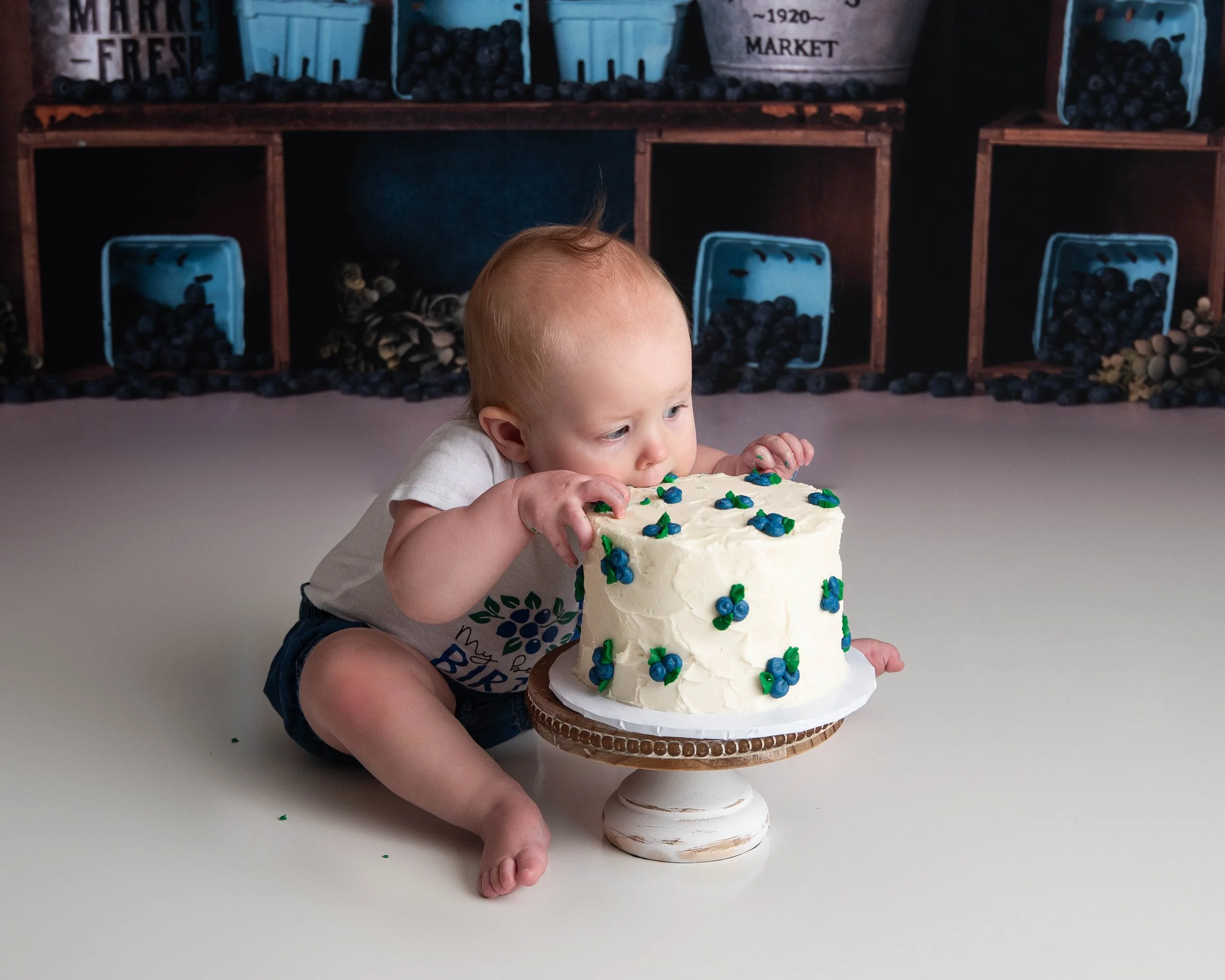A baby sitting on the floor and about to blow out birthday candles on a white frosted cake decorated with blue and green flowers, placed on a cake stand.