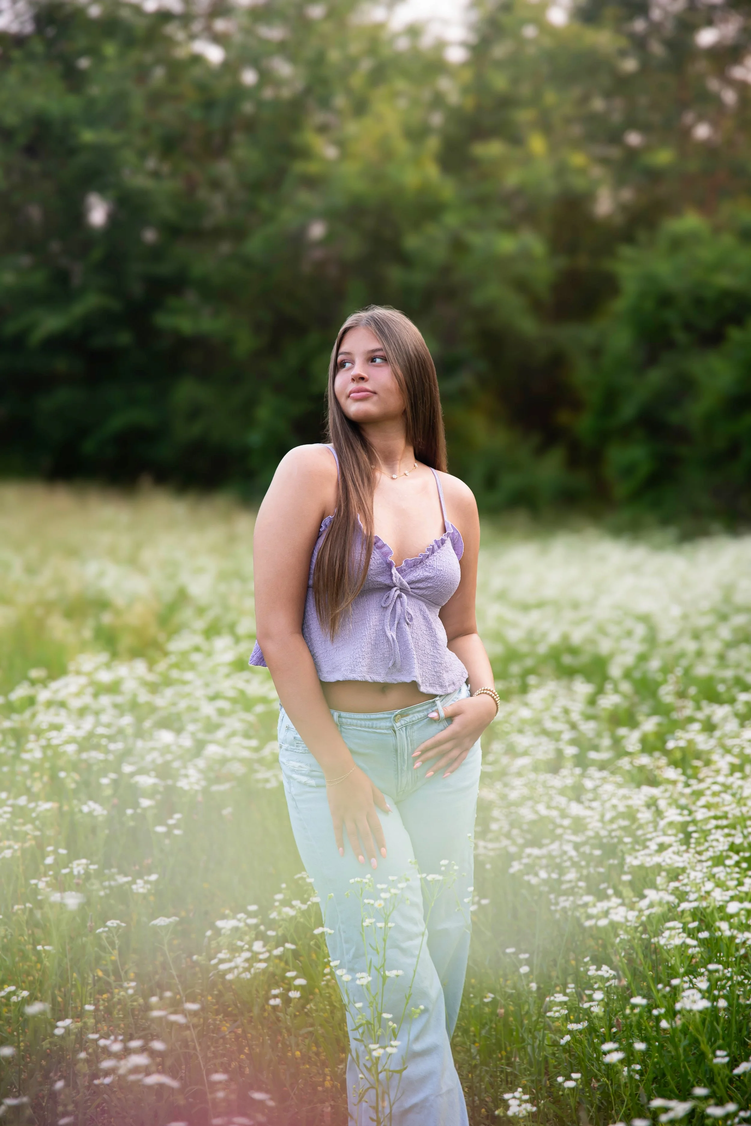 A young woman standing in a field of white flowers with green trees in the background, wearing a purple top and light blue jeans.
