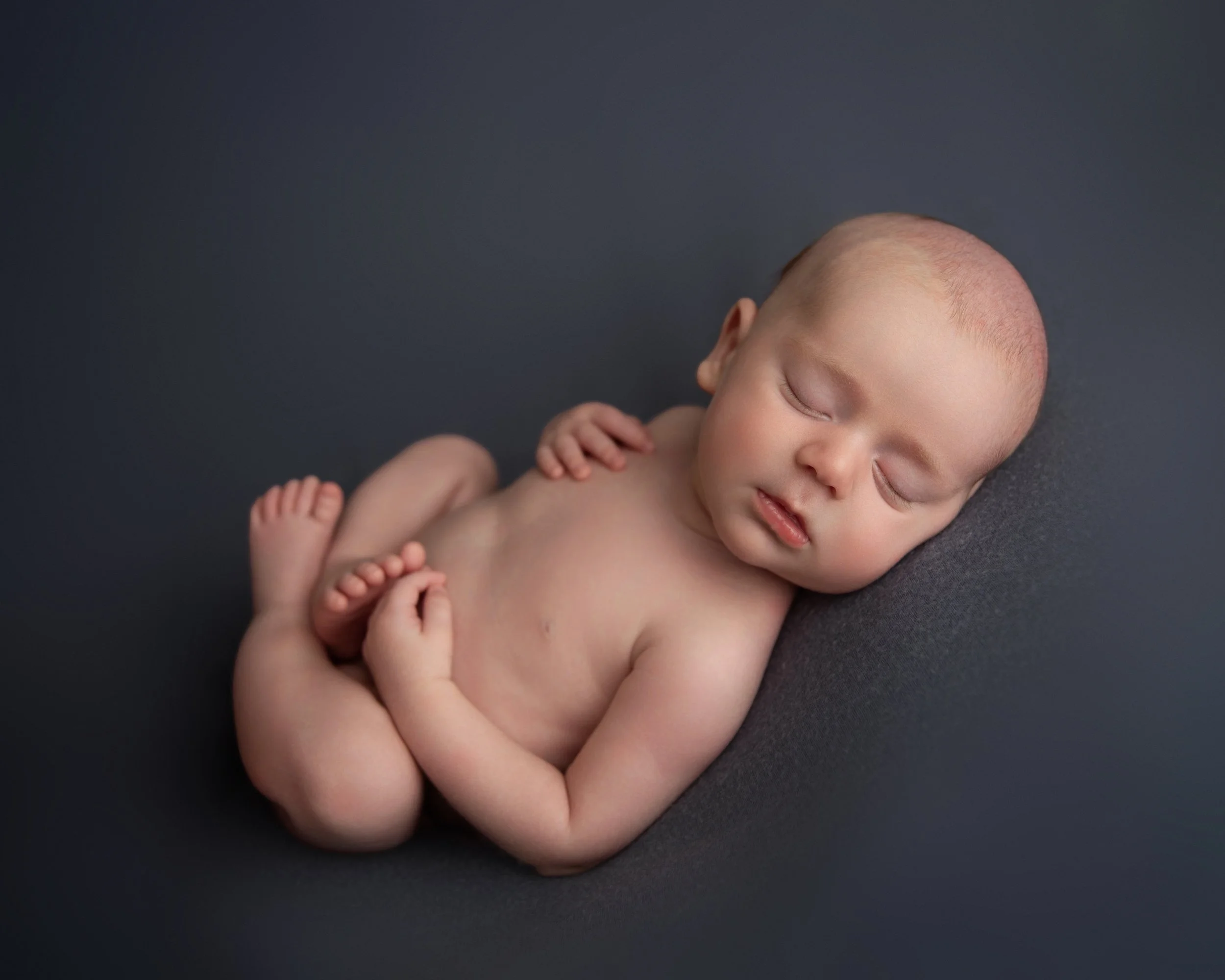 A sleeping baby lying on a dark surface, with arms and legs curled up, and eyes closed.