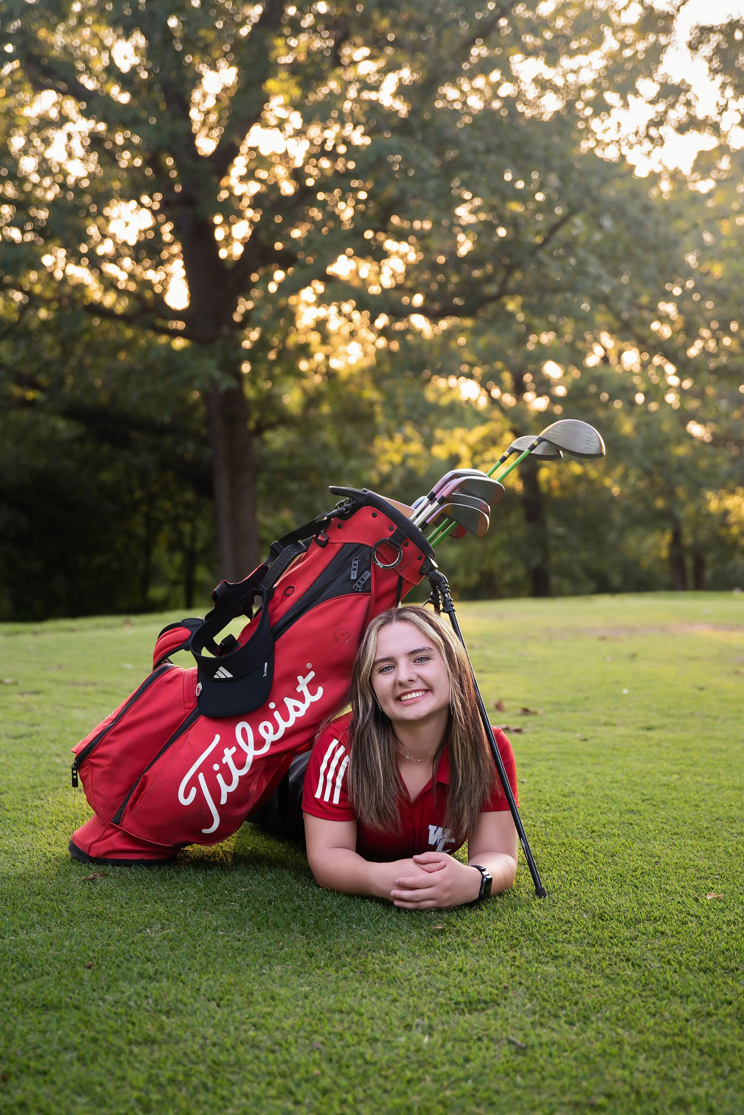 A young woman lying on a golf course with a red golf bag and clubs, smiling at the camera during sunset.