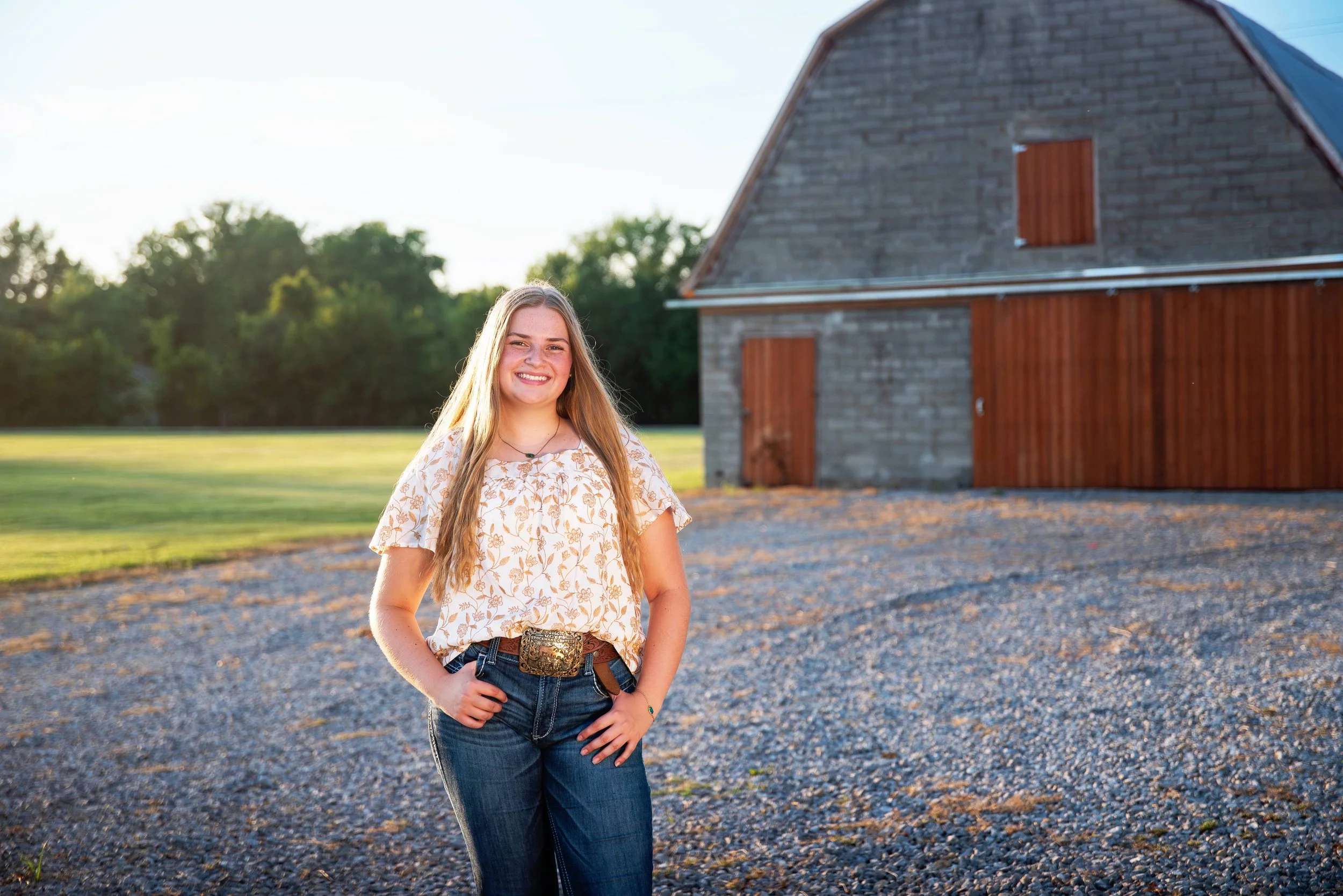 Young woman standing outdoors near a barn during sunset, smiling with hand on her hip.