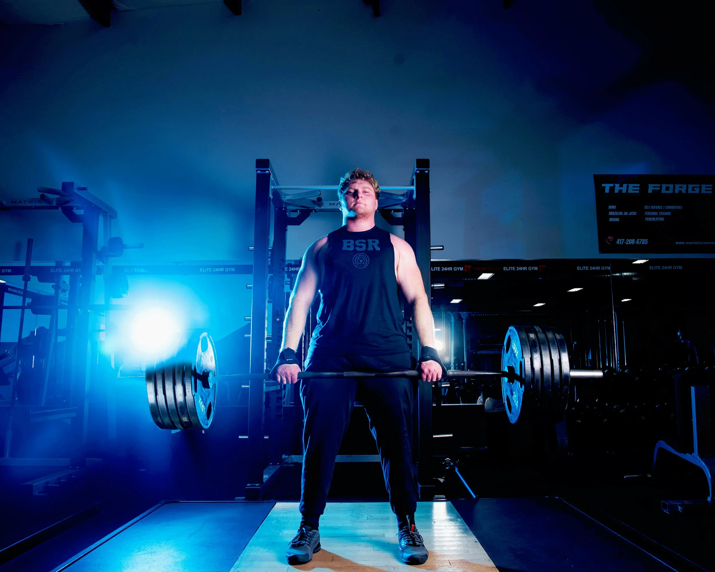 A young man lifting a barbell with weights in a gym, standing in front of a squat rack with blue lighting and a large digital screen on the right side.