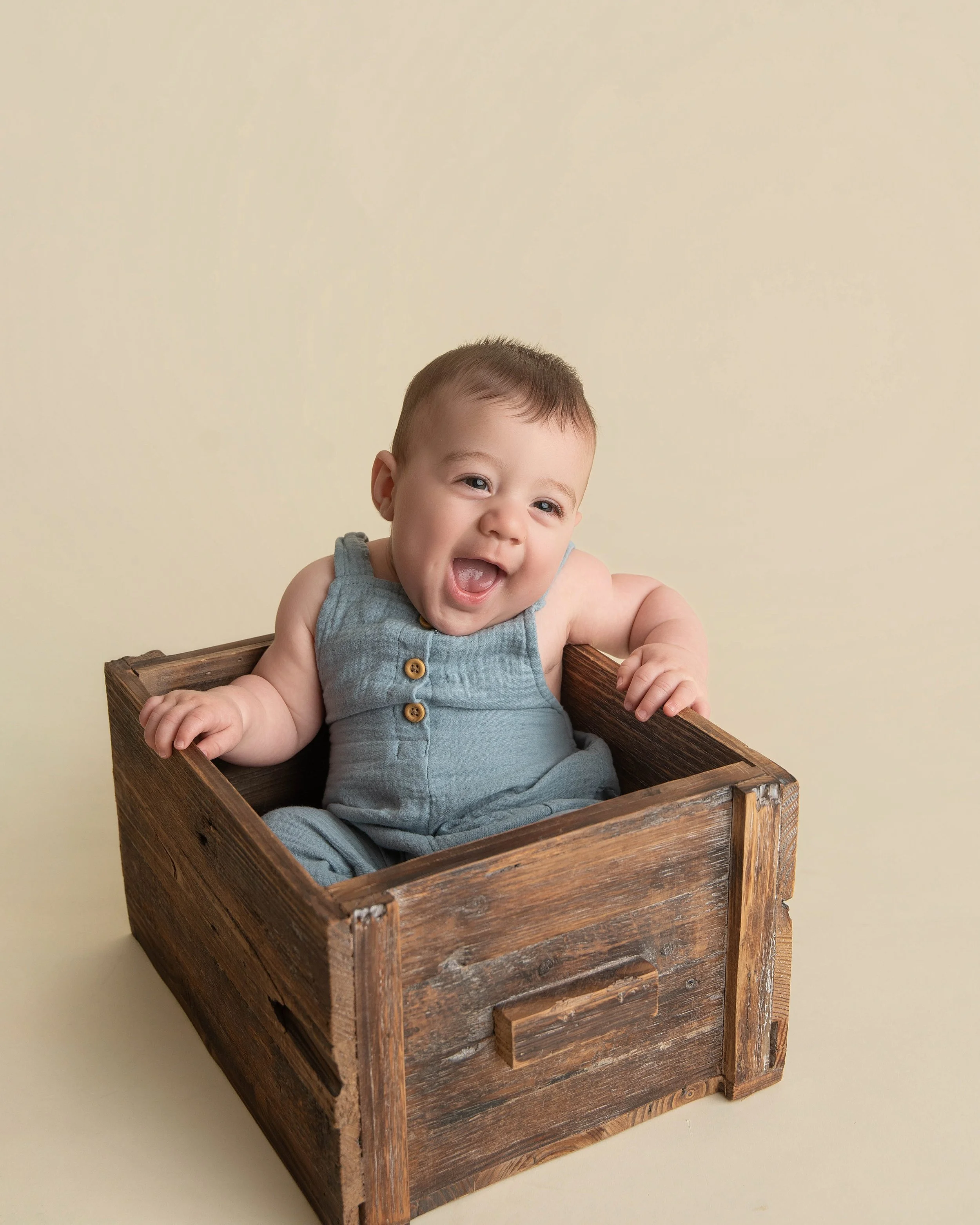 A smiling baby sitting inside a wooden crate against a light beige background.