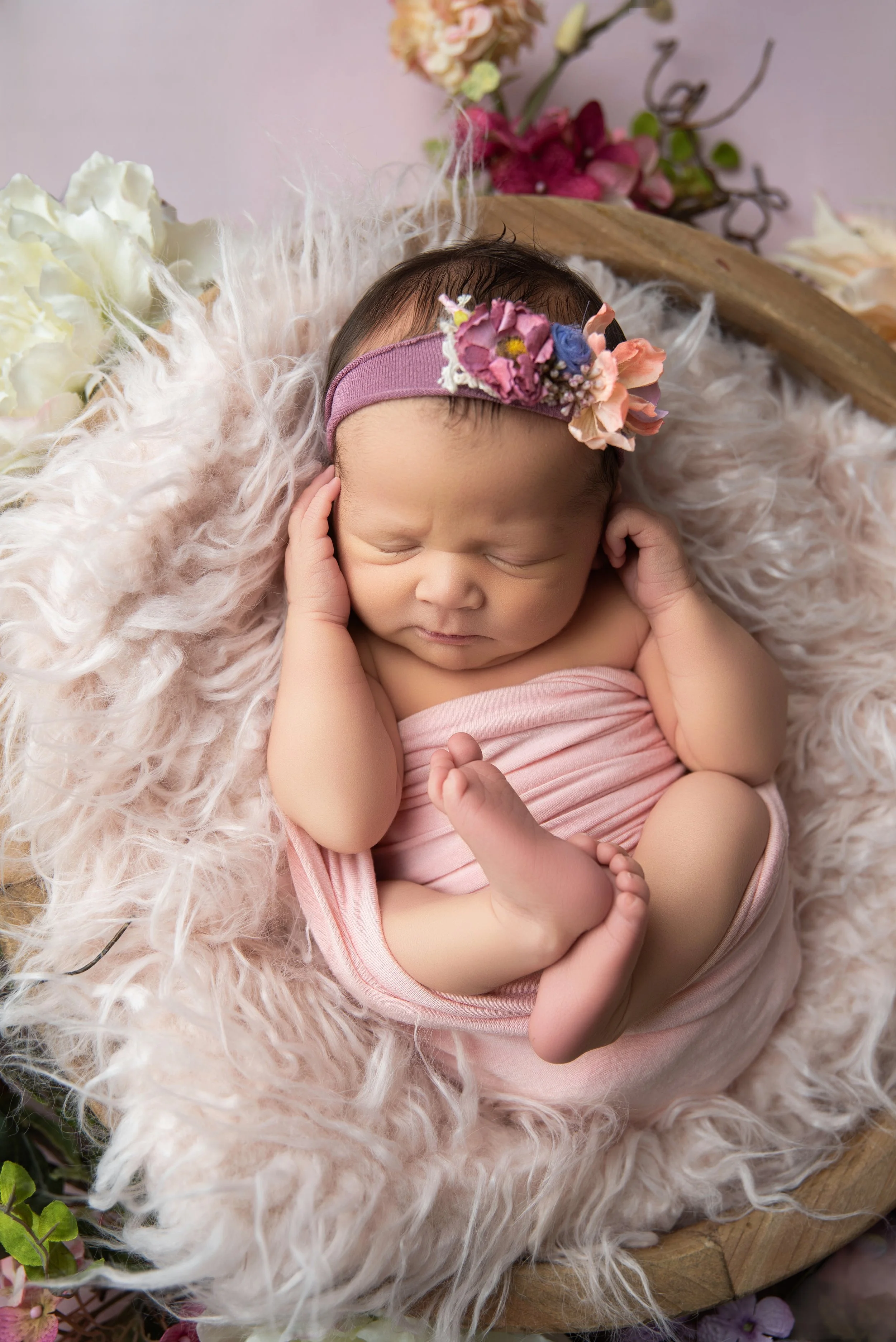 A sleeping newborn baby girl wrapped in pink cloth, lying on a fluffy pink blanket inside a wooden basket, wearing a pink floral headband.