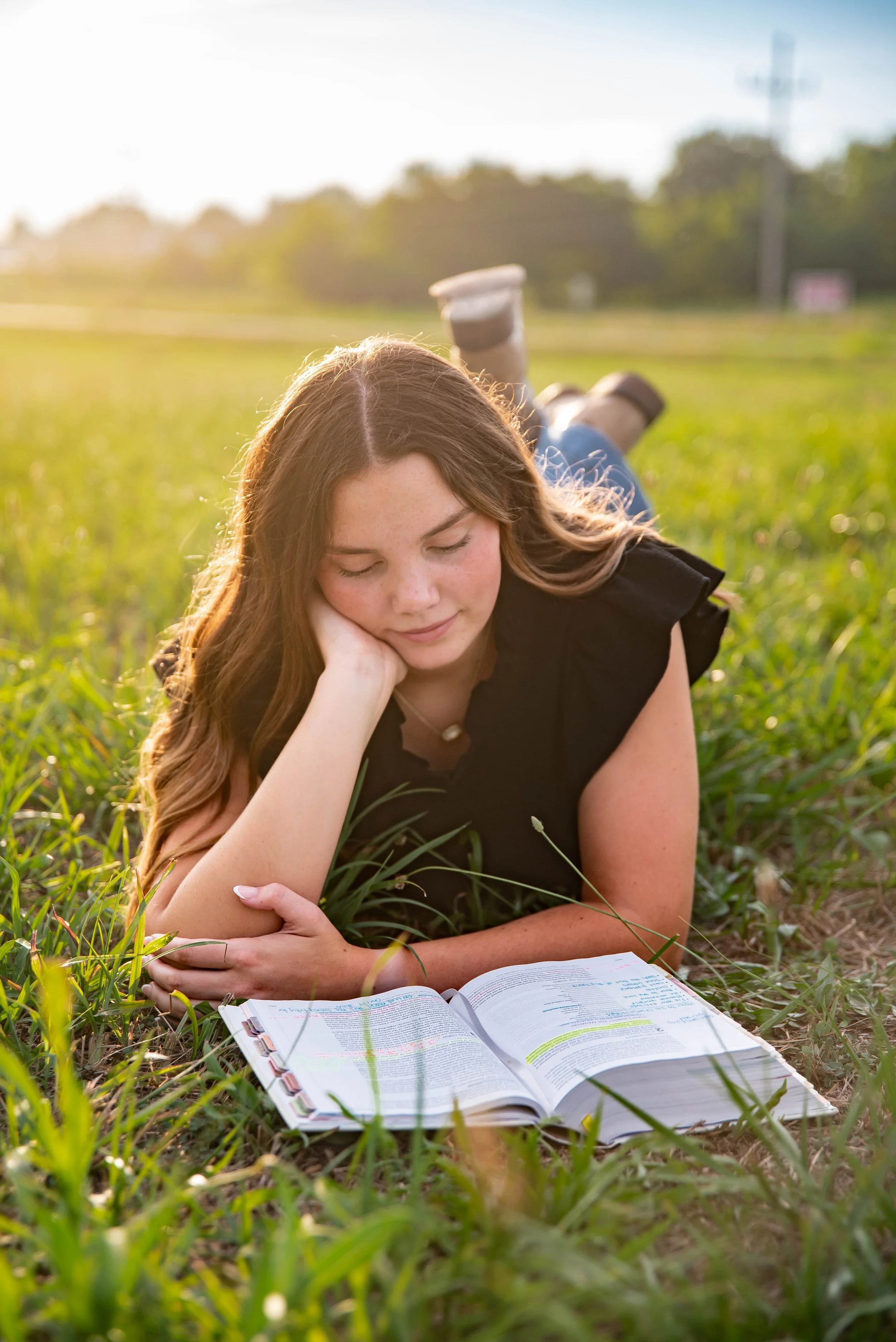 A young girl with long, wavy brown hair lying on her stomach in a grassy field, reading a large open book with handwritten notes, during sunset.