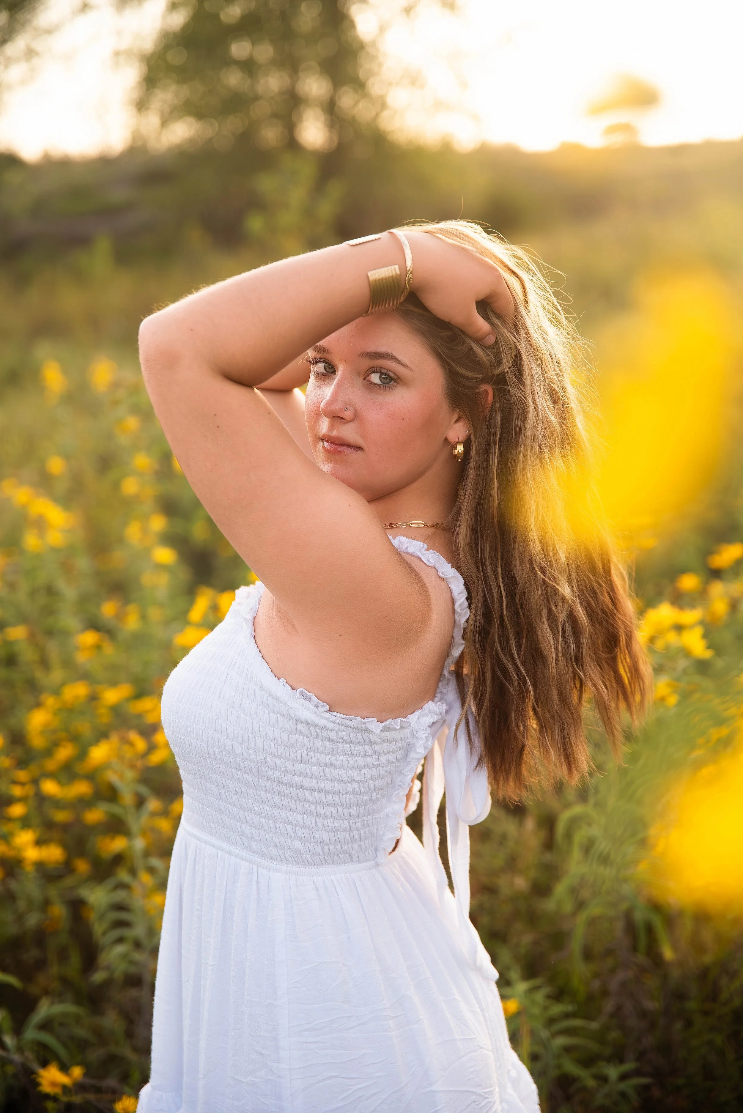 A woman with long wavy hair, wearing a white dress, standing in a field of yellow flowers during sunset, posing with one arm above her head.