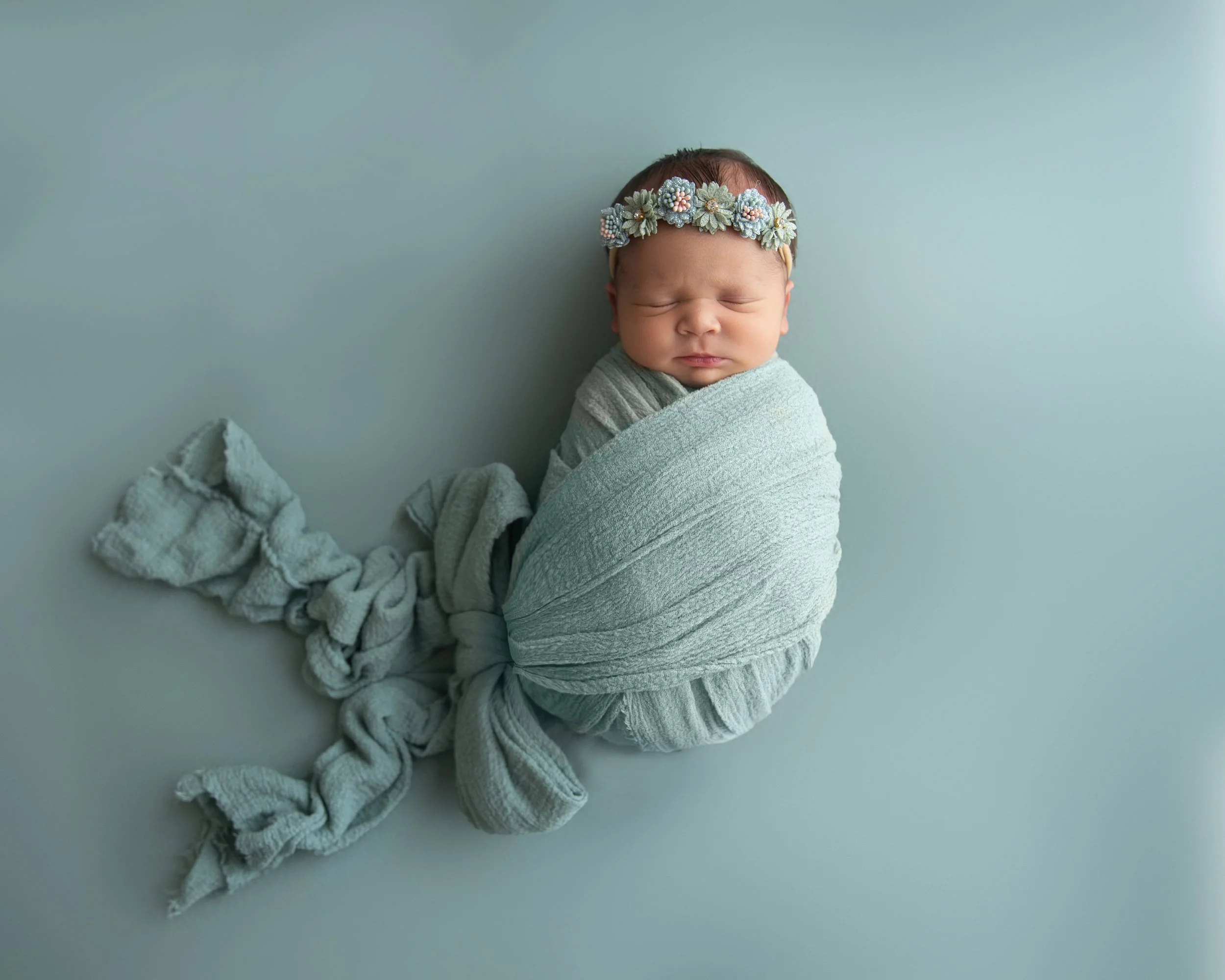 Newborn baby wrapped in a light grey blanket, wearing a floral headband, sleeping against a plain light background.