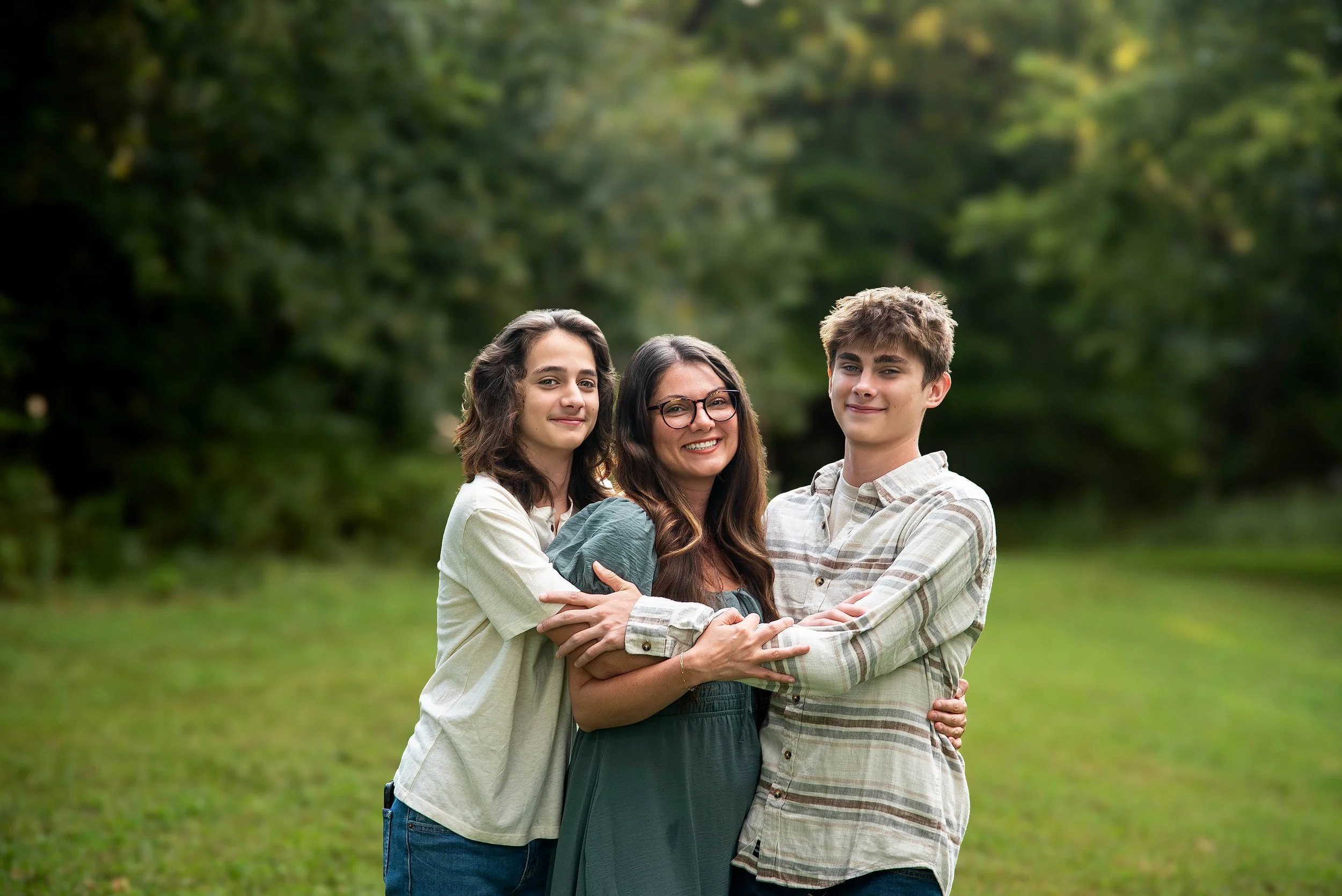 Three people, a woman and two teenagers, standing together outdoors in a grassy area with trees, smiling and hugging each other.