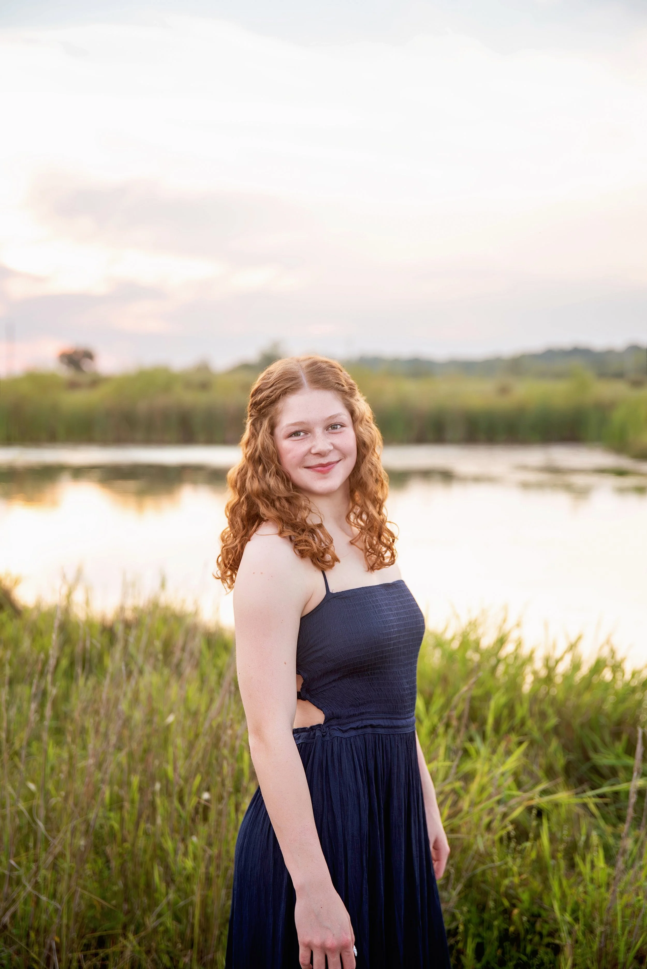 A young woman with curly red hair in a navy dress standing near a pond during sunset.