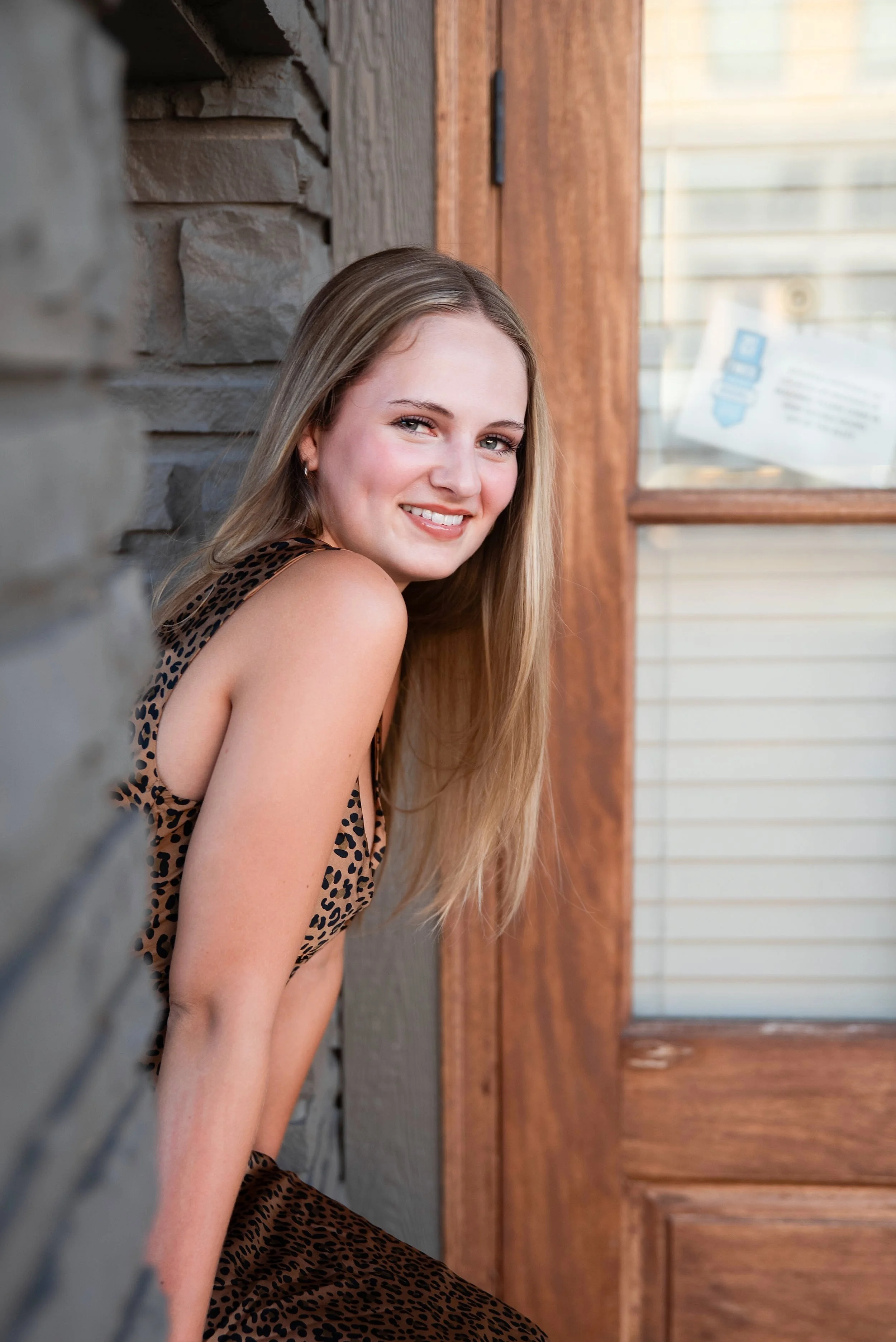 A young woman with long blonde hair smiling and leaning against a gray stone wall outside a wooden building with a glass window.