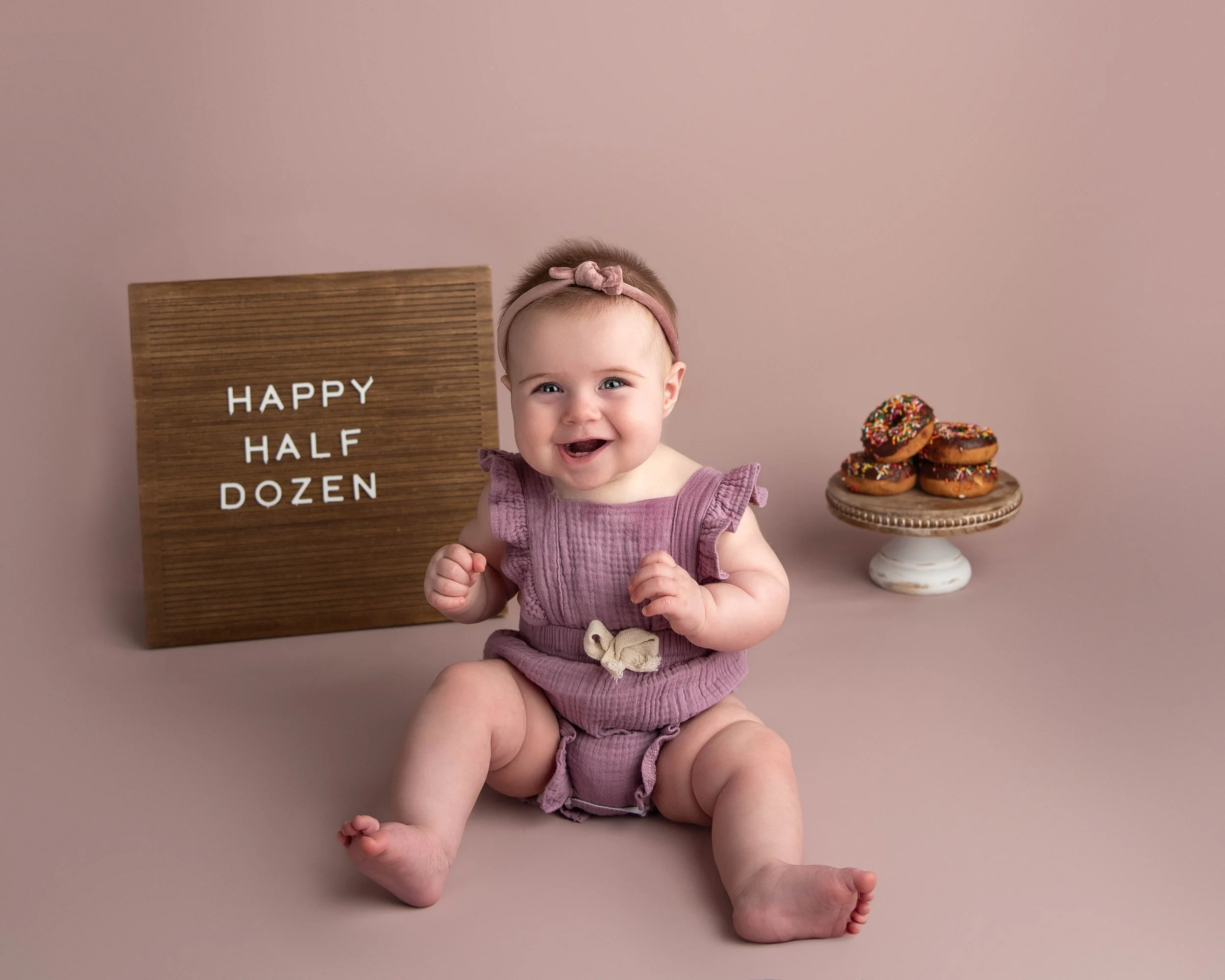 A smiling baby girl sitting on the floor with a light pink background, a wooden sign that reads 'HAPPY HALF DOZEN,' and a plate of donuts with colorful sprinkles.