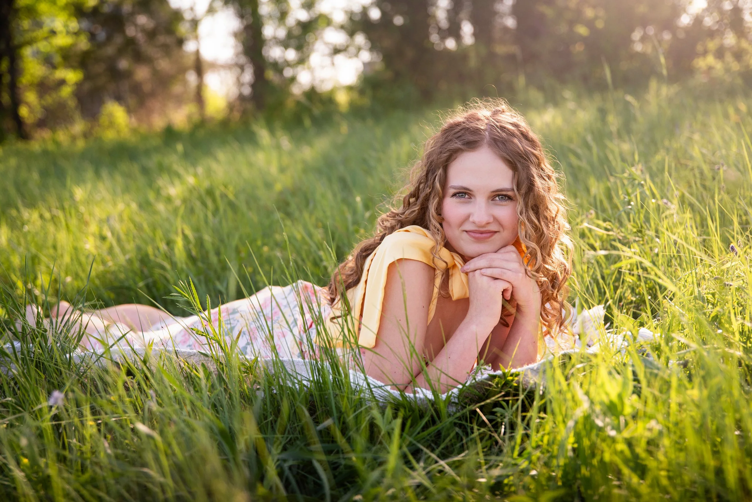 A young woman with curly blonde hair lying on her stomach in a grassy field, resting her chin on her hands, looking at the camera with a slight smile, with sunlight filtering through trees in the background.