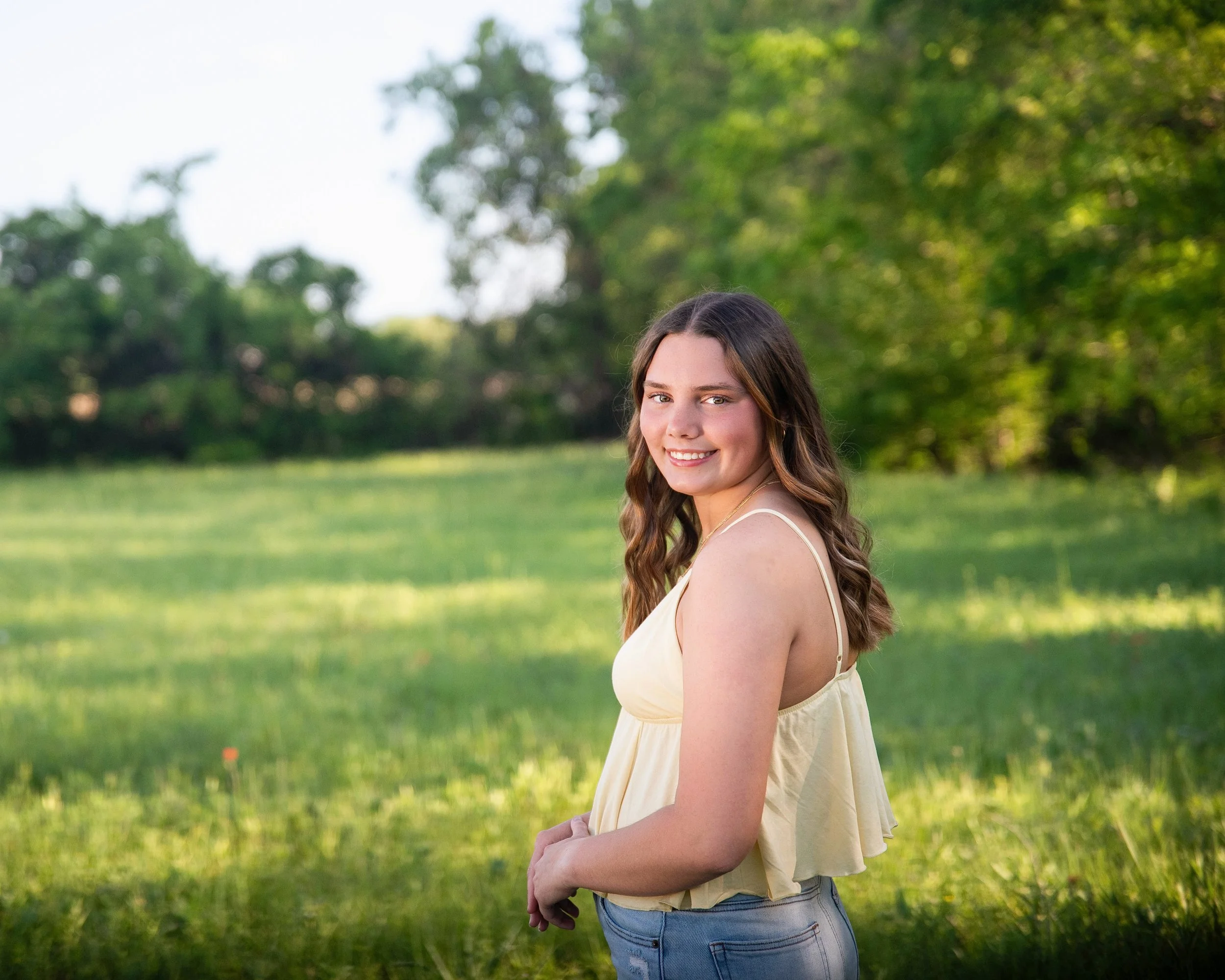 Young woman with wavy brown hair and a beige summer top standing in a green outdoor field during daytime.