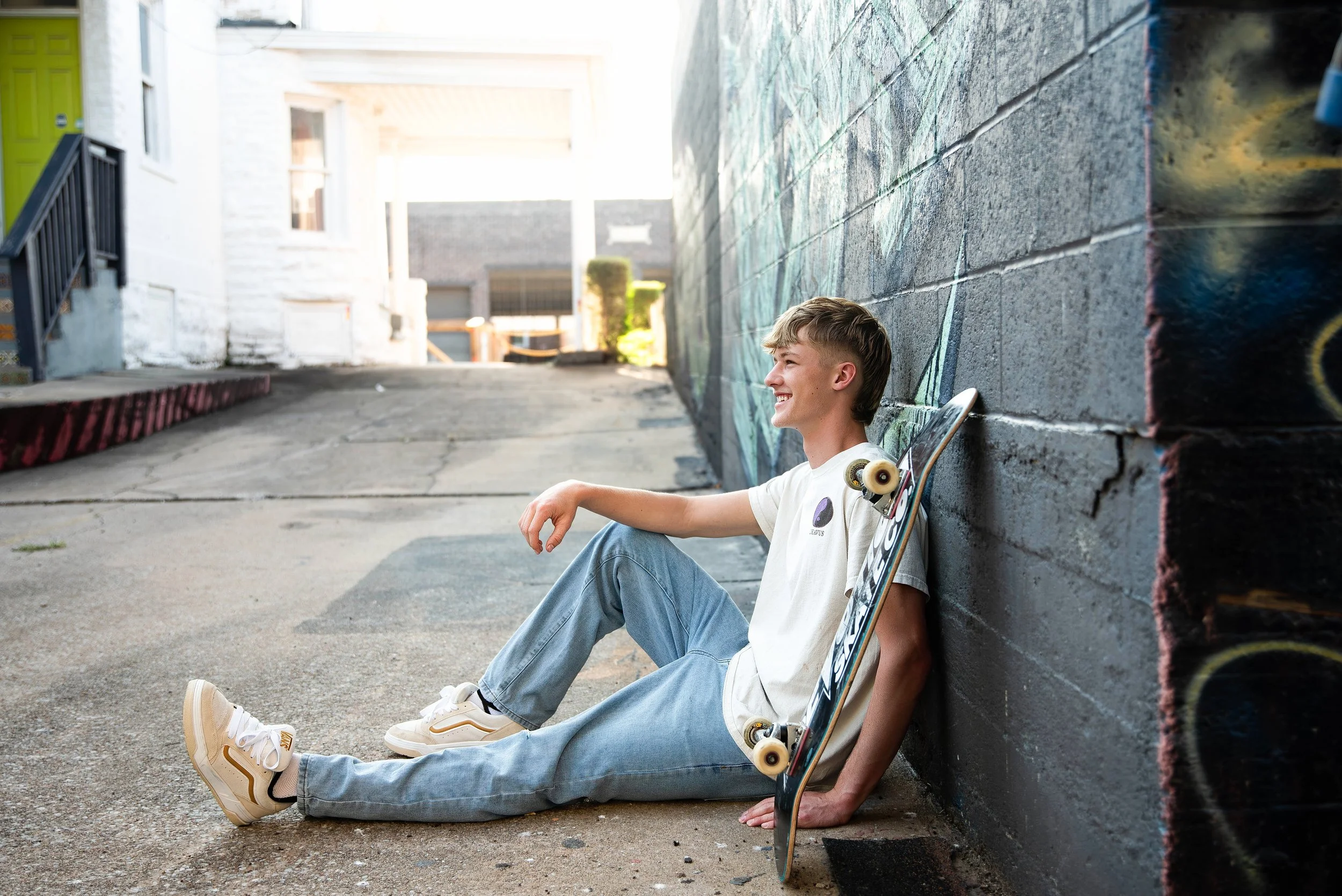 A young man with a skateboard sitting on the ground and leaning against a graffiti-covered brick wall in an alley.