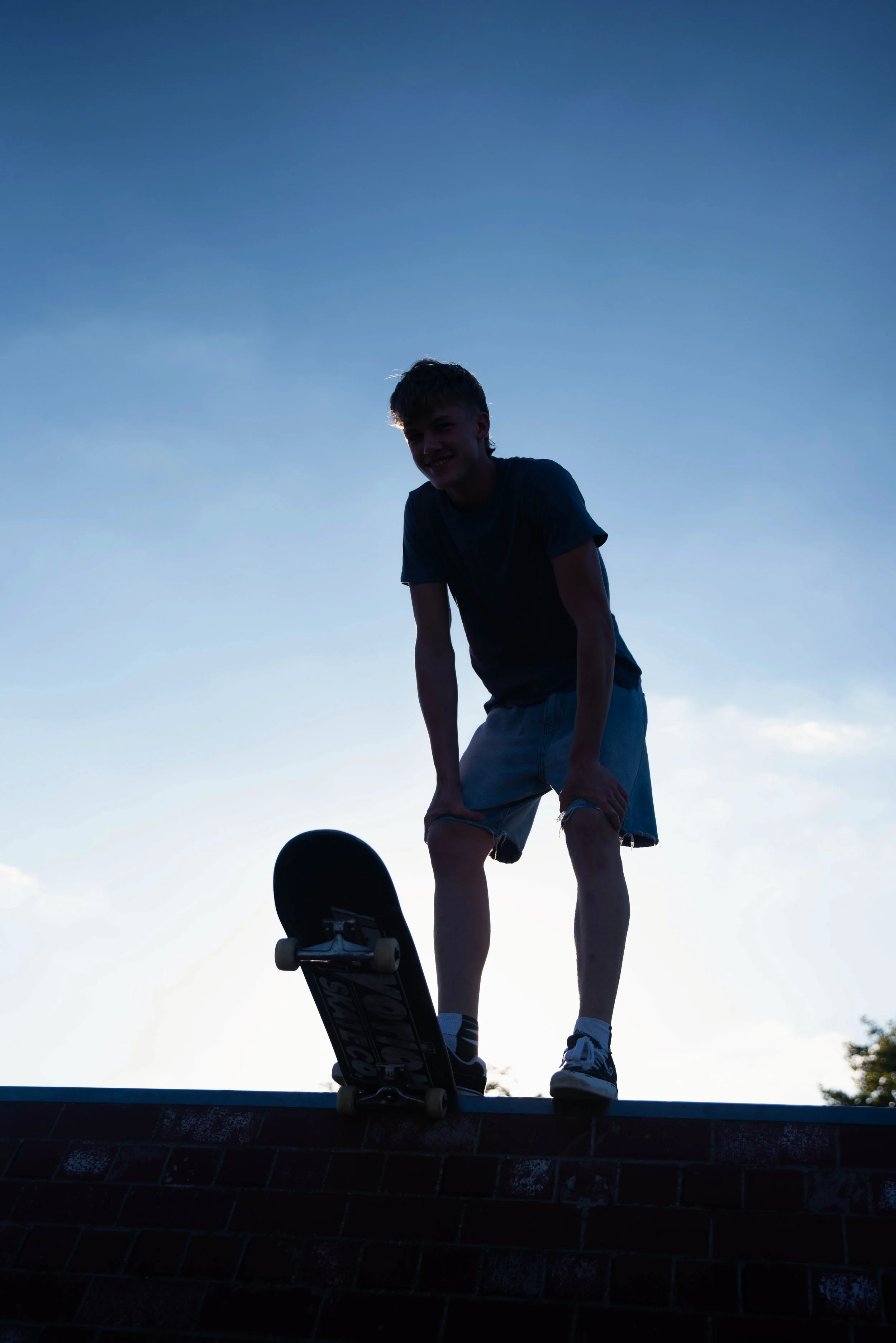 A silhouette of a boy with a skateboard, standing on a brick ledge against the sky.