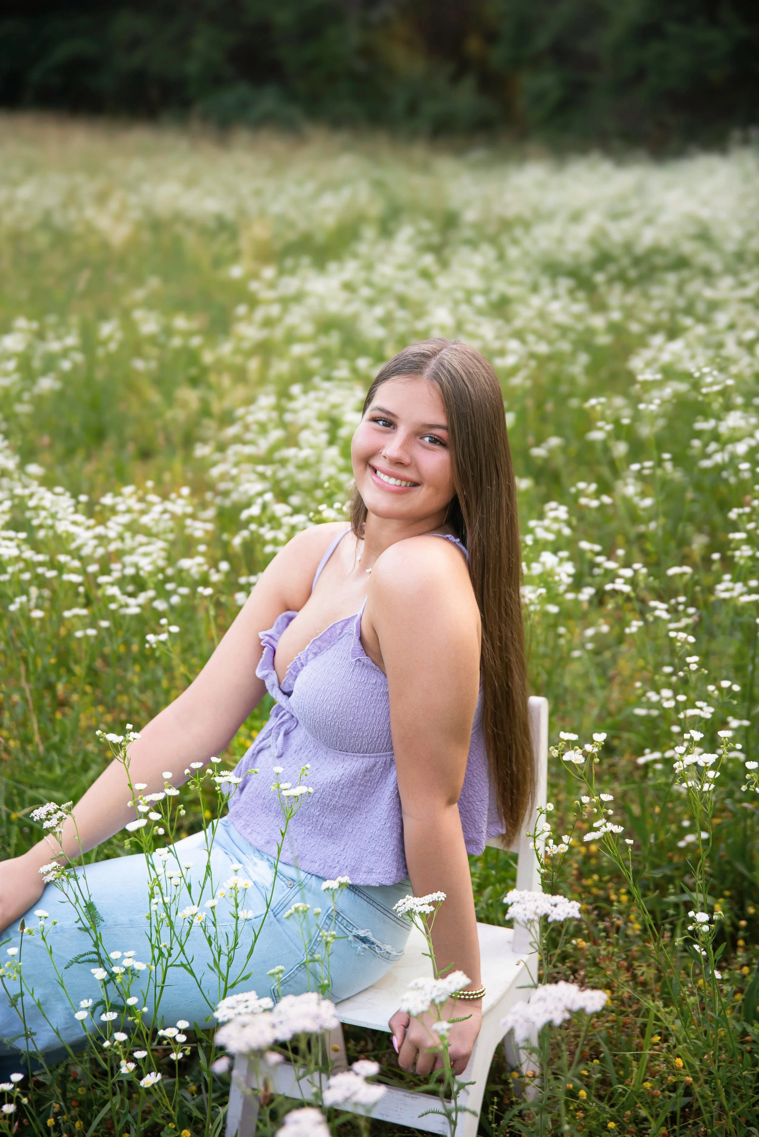 A young woman with long brown hair sitting on a white bench in a field of white wildflowers, smiling at the camera, wearing a lavender sleeveless top and light blue jeans.