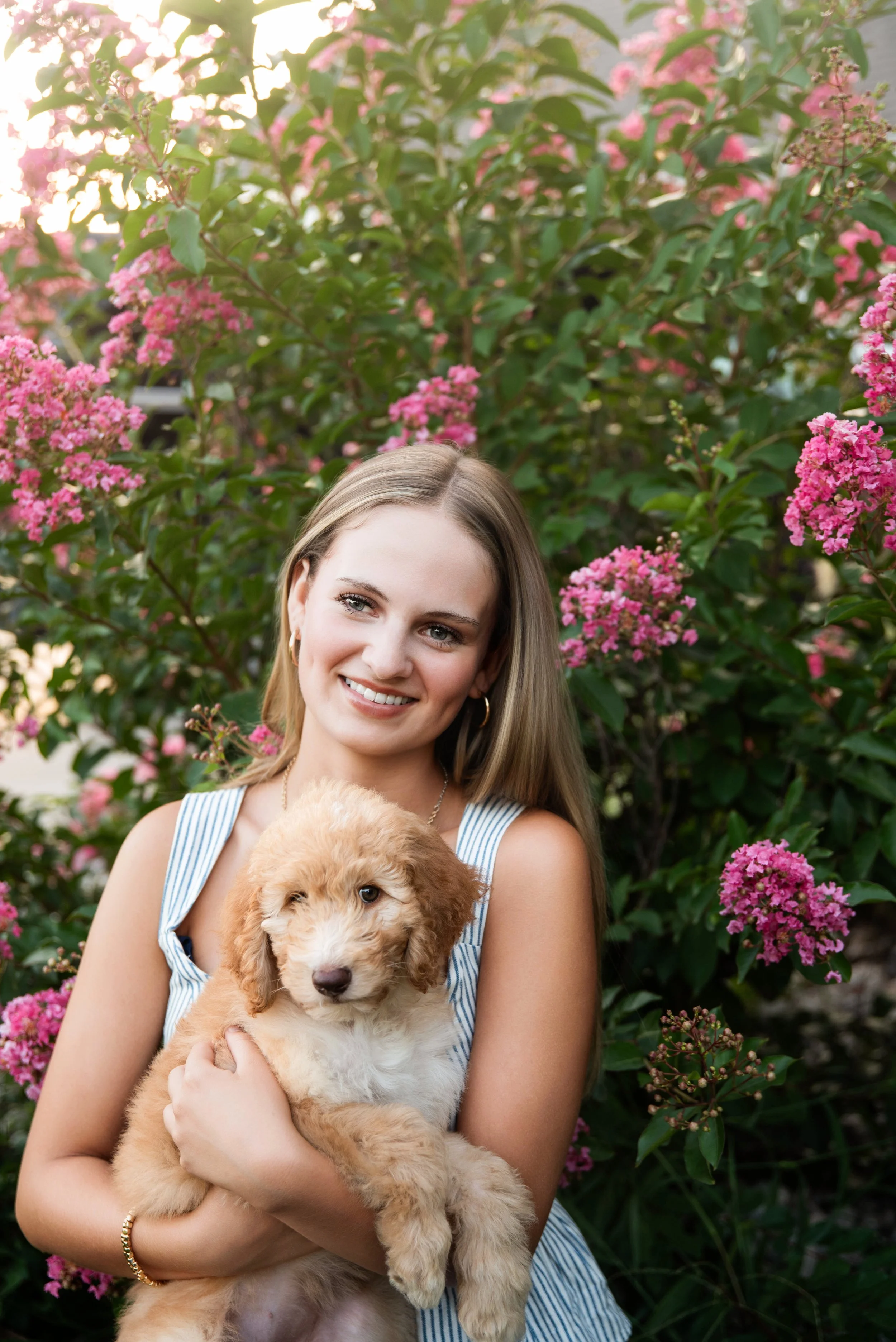 A young woman with long, light brown hair, wearing a sleeveless blue and white striped dress and gold hoop earrings, holding a fluffy, light brown puppy with white markings, surrounded by pink flowering bushes in a garden.