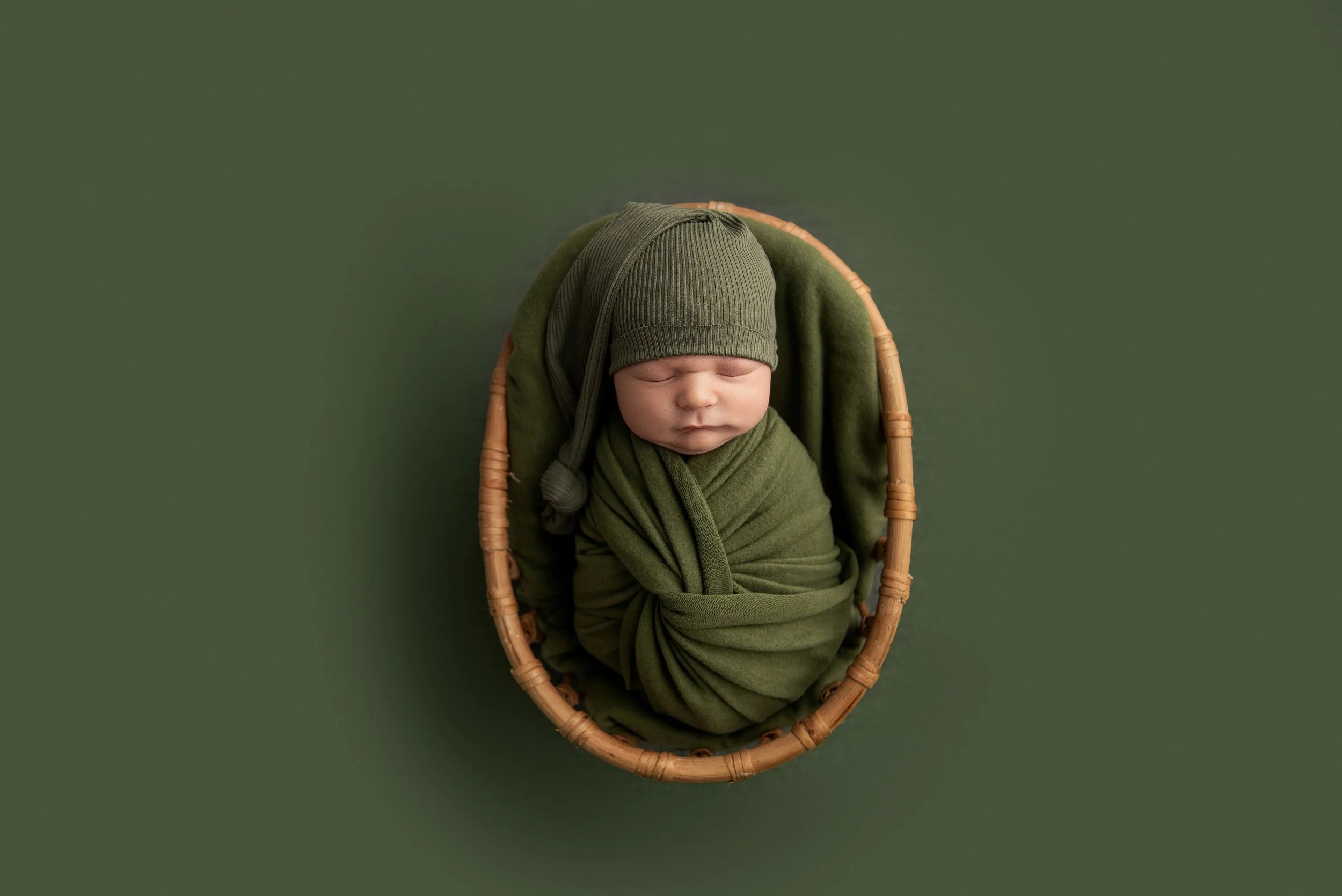 A newborn baby wrapped in green cloth and wearing a matching knitted hat, sleeping in a wicker basket on a green background.