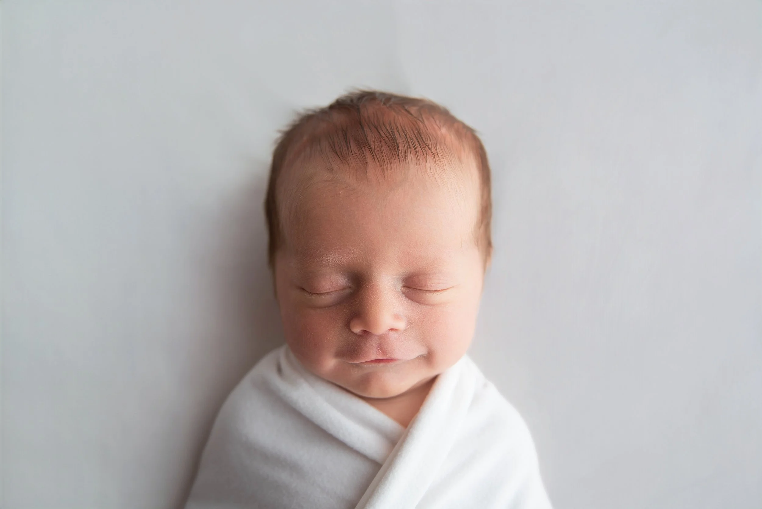 Close-up of a peaceful newborn baby wrapped in a white blanket with eyes closed and slight smile, soft lighting, plain light gray background.