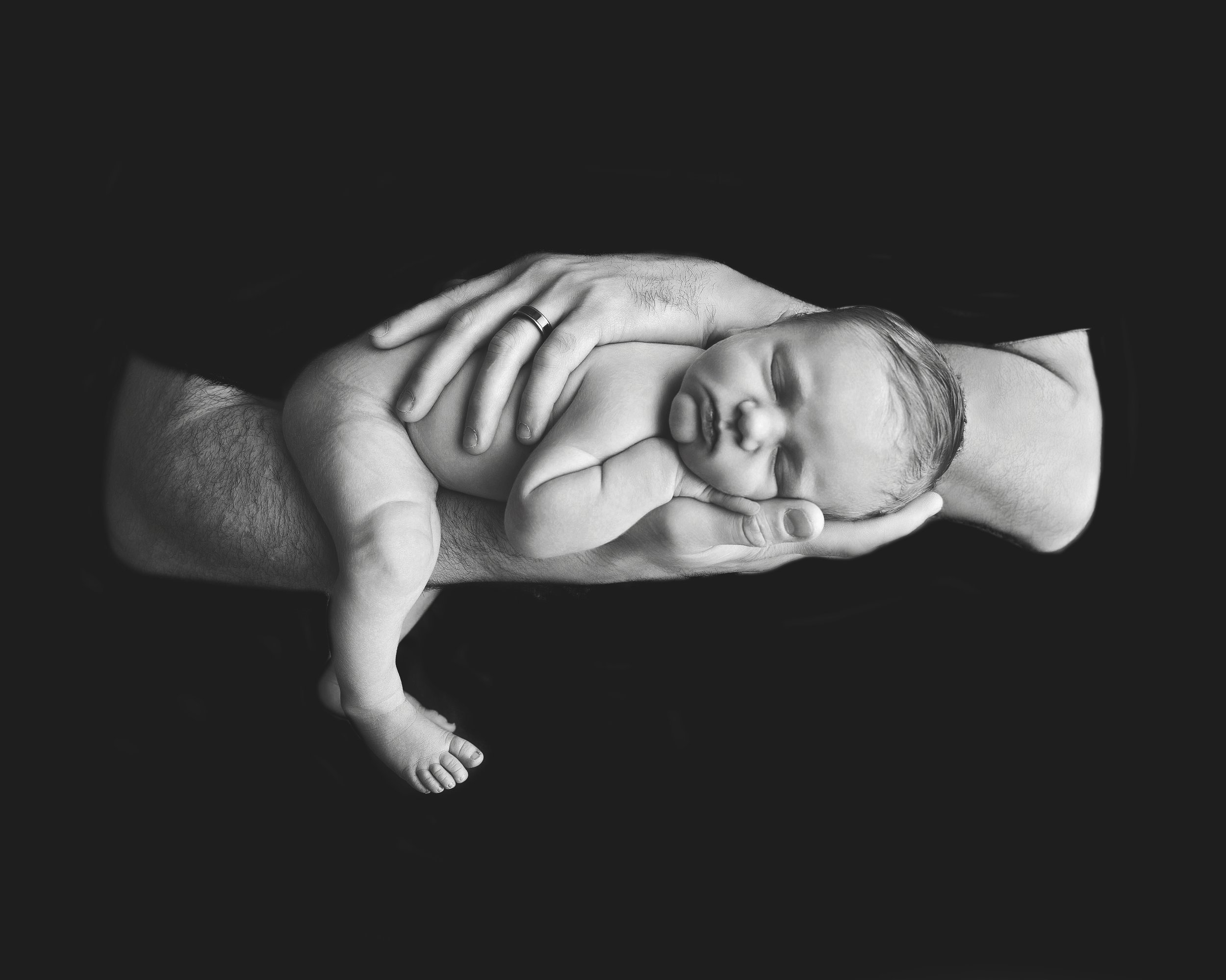 Black and white photo of a newborn baby being held gently in adult hands, resting on a dark background.
