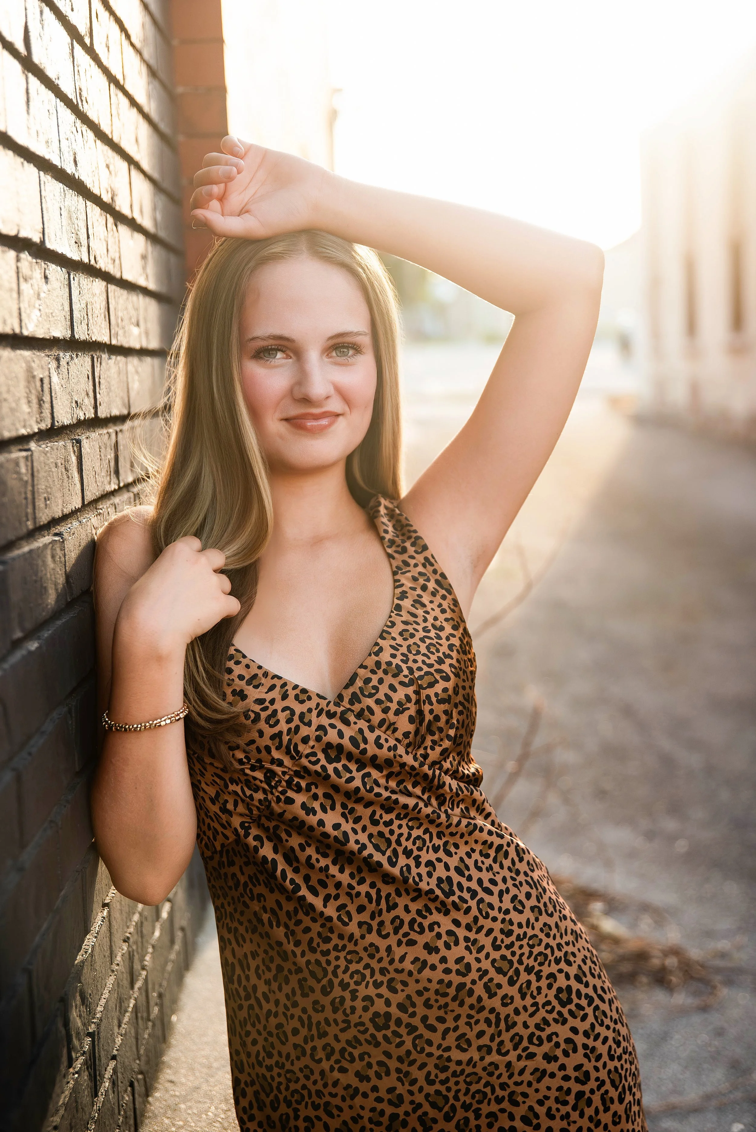 A young woman with long brown hair wearing a leopard print dress, standing against a brick wall in sunlight, with one arm raised above her head and the other hand on her shoulder, smiling gently at the camera.