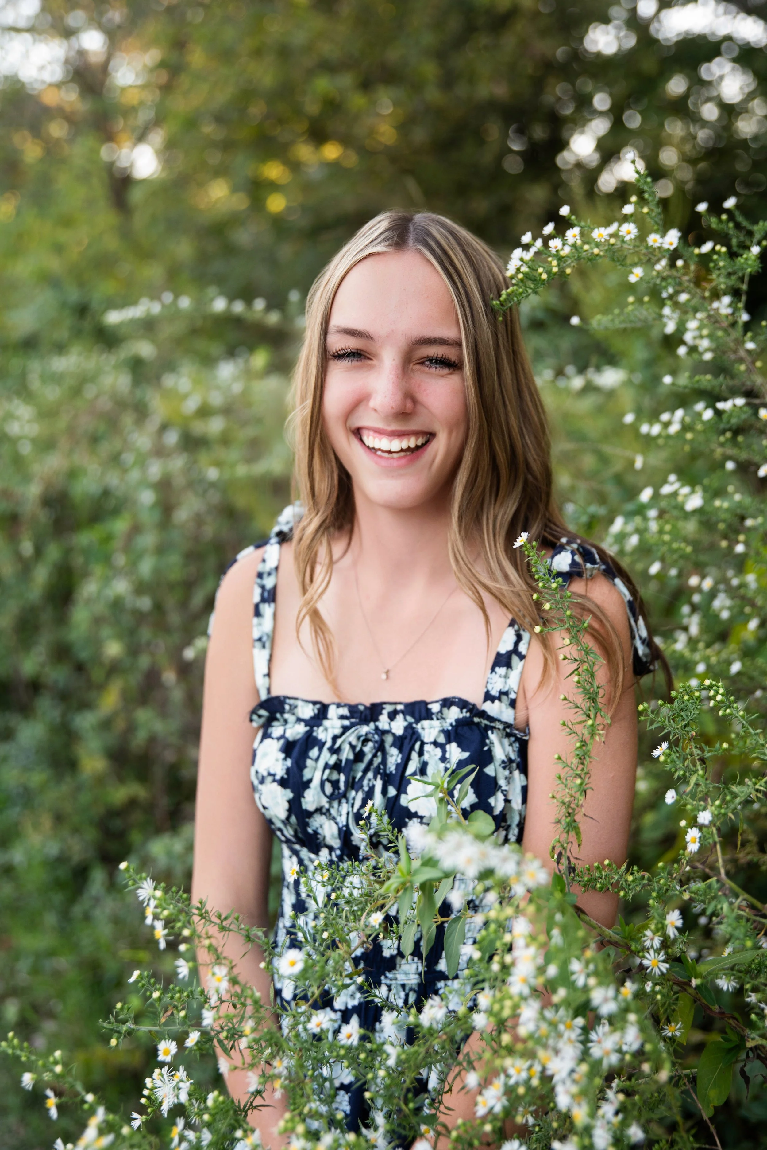 A young woman with long, wavy, light brown hair and blue eyes, wearing a navy blue dress with white floral patterns, standing outdoors surrounded by greenery and small white flowers, smiling broadly.