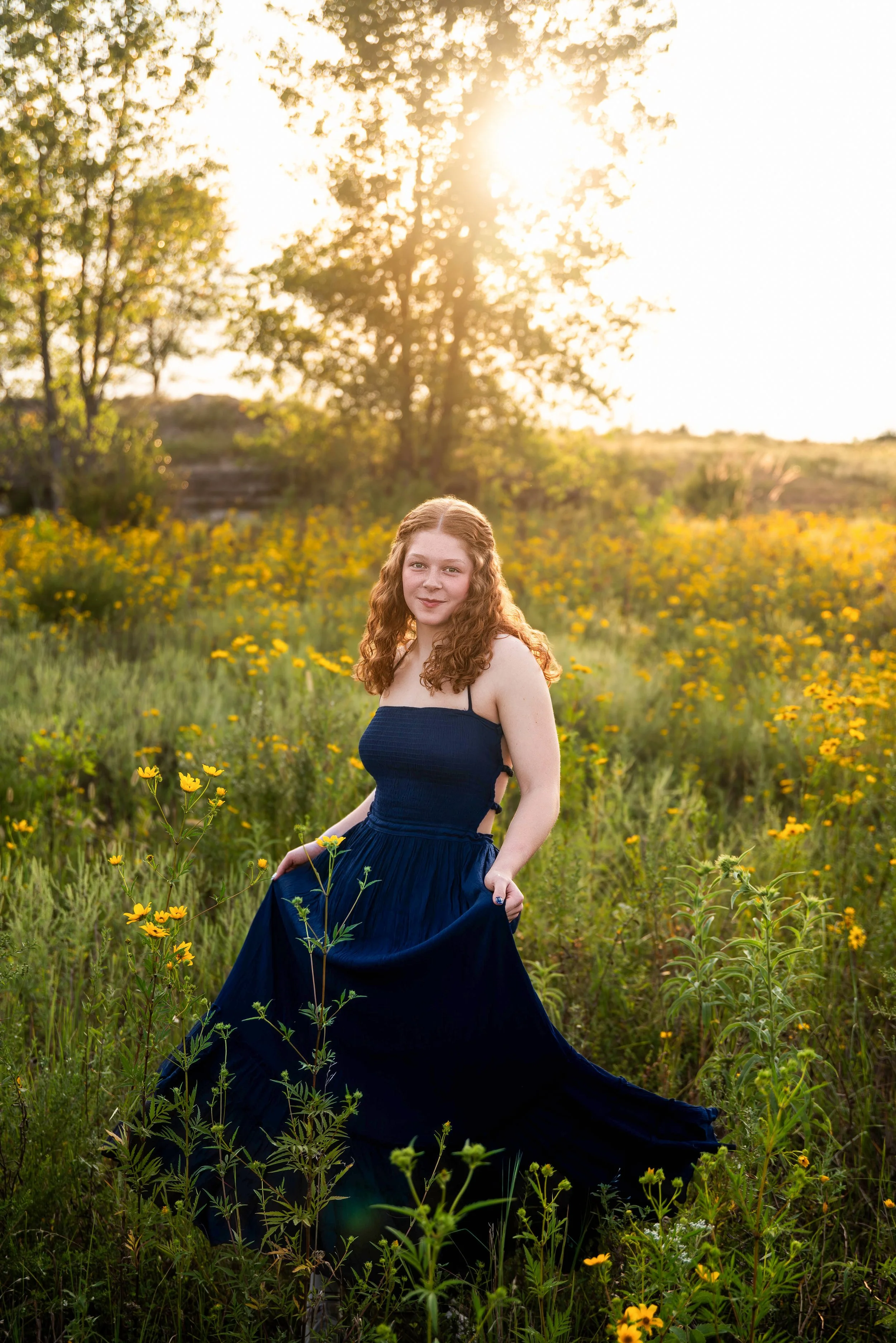 A young woman with curly red hair standing in a field of yellow flowers during sunset, wearing a dark blue dress.