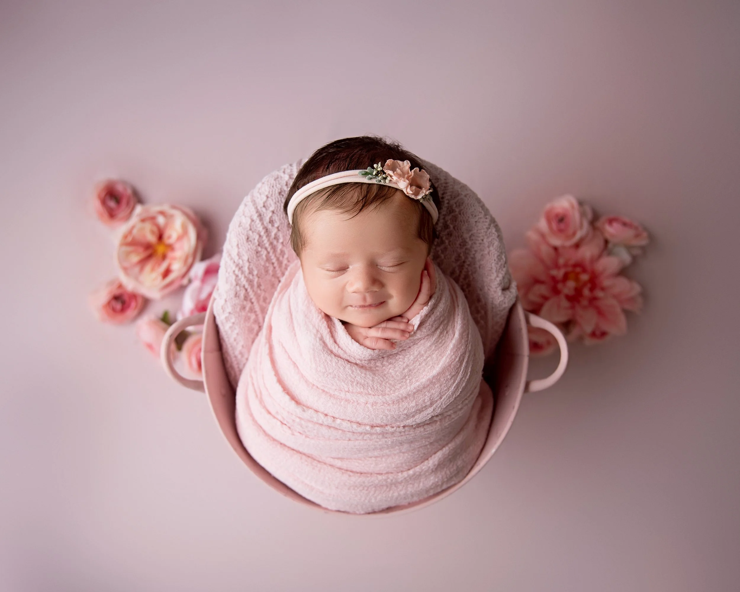 A newborn baby wrapped in a pink blanket, wearing a floral headband, and lying in a pink basket surrounded by pink flowers on a pink background.