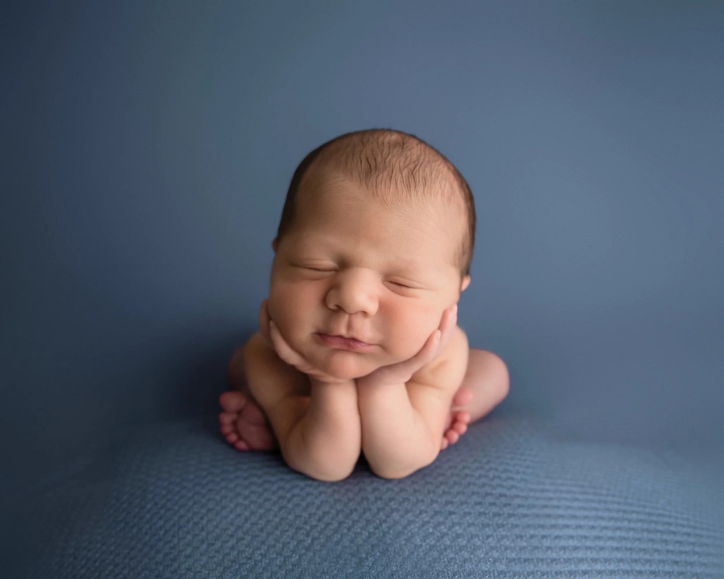 A newborn baby with closed eyes, resting head on hands, and smiling face, against a blue background.