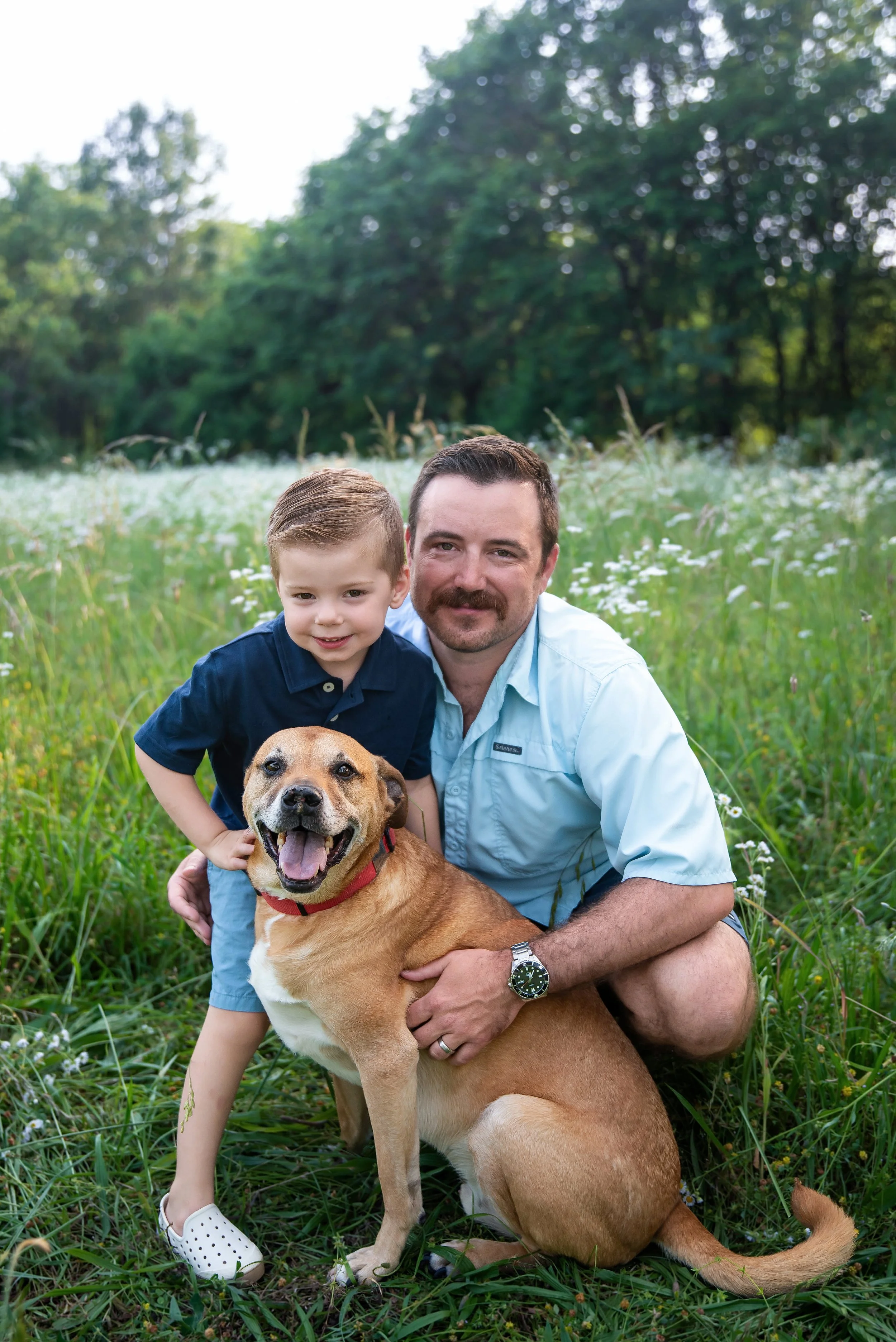 A man, a young boy, and a dog outdoors in a grassy field with trees in the background.