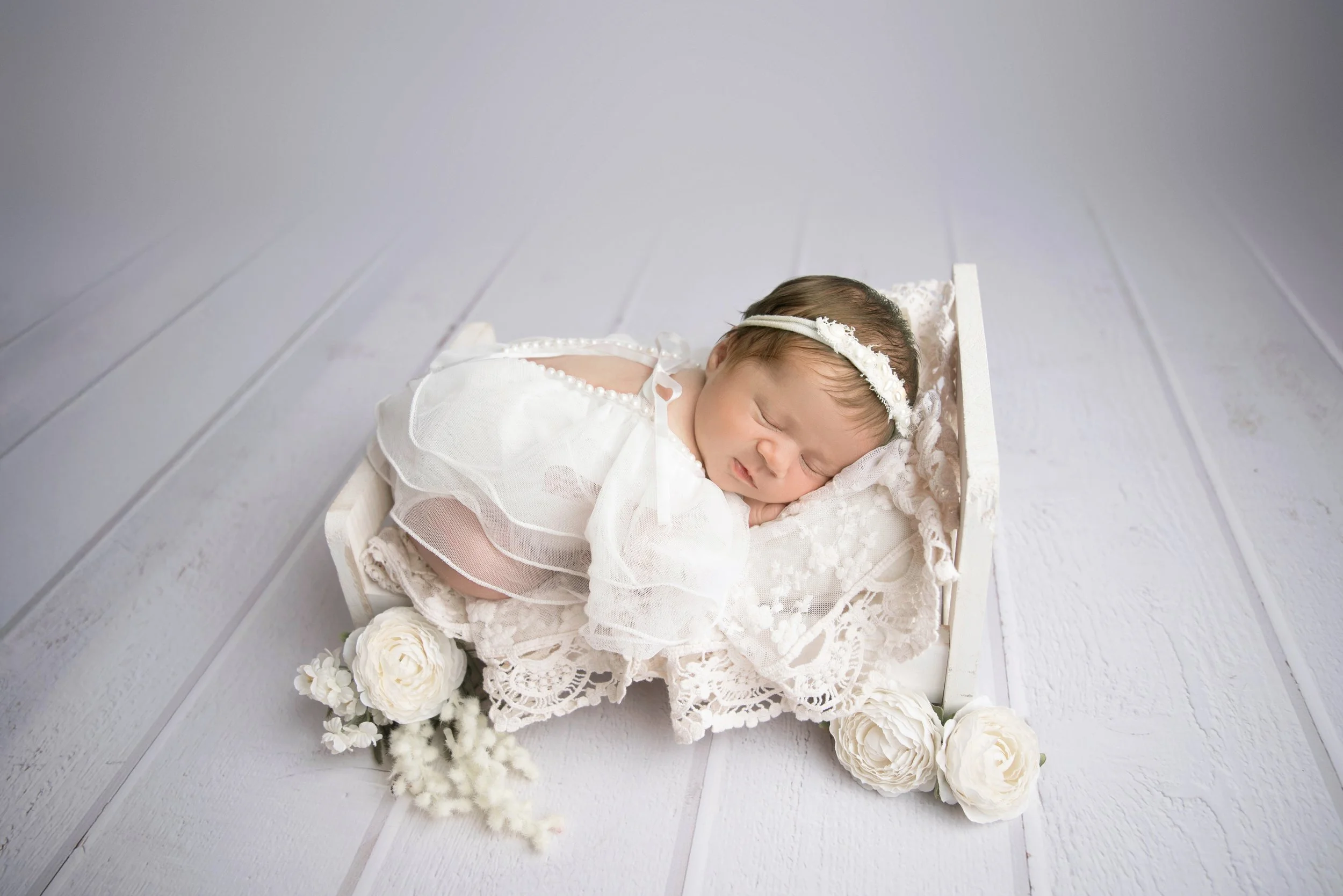 A sleeping baby girl dressed in white lace, lying on a lace blanket in a small white bed with white flowers and soft fabric decorations.