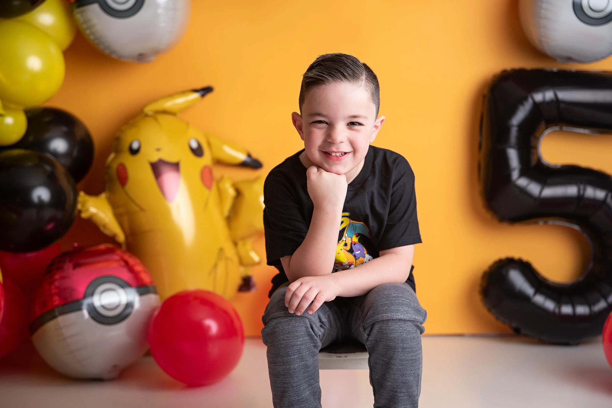 Smiling young boy with short dark hair, wearing a black Pokémon T-shirt and gray pants, sitting on a chair with his chin resting on his hand, surrounded by Pokémon themed balloons including Pikachu, Pokéballs, and a large black number 3 balloon, agai