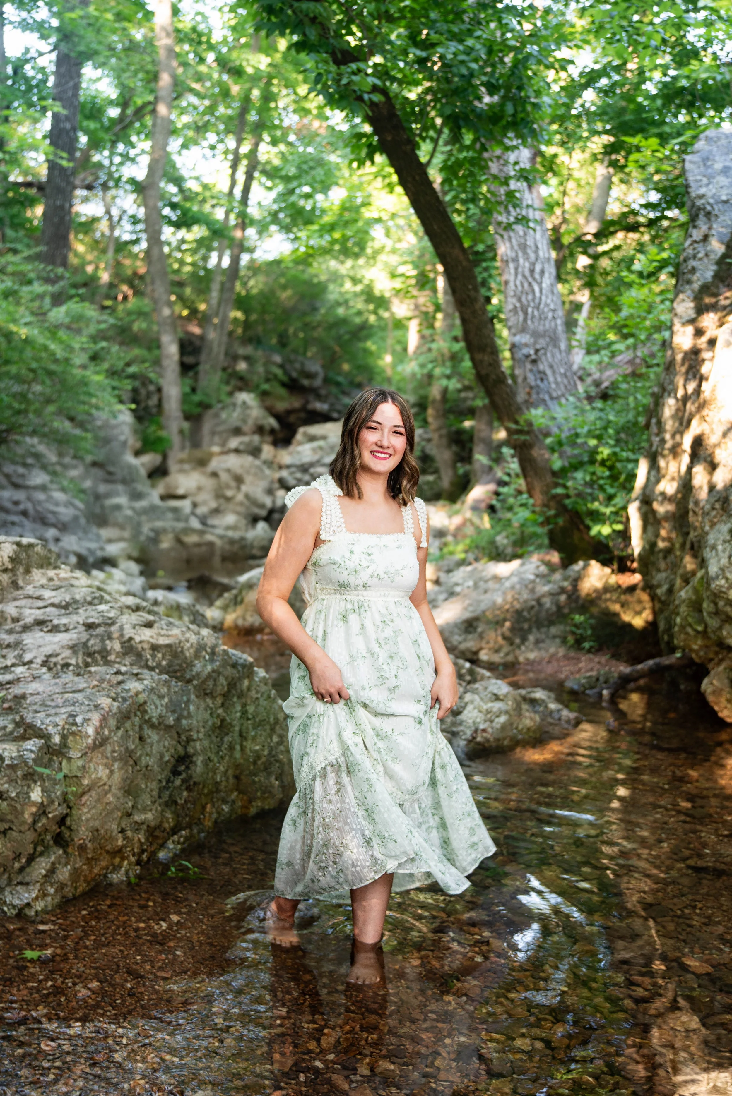 A young woman wearing a white dress with green floral patterns, standing barefoot in a shallow creek surrounded by rocks and lush green trees, smiling at the camera.