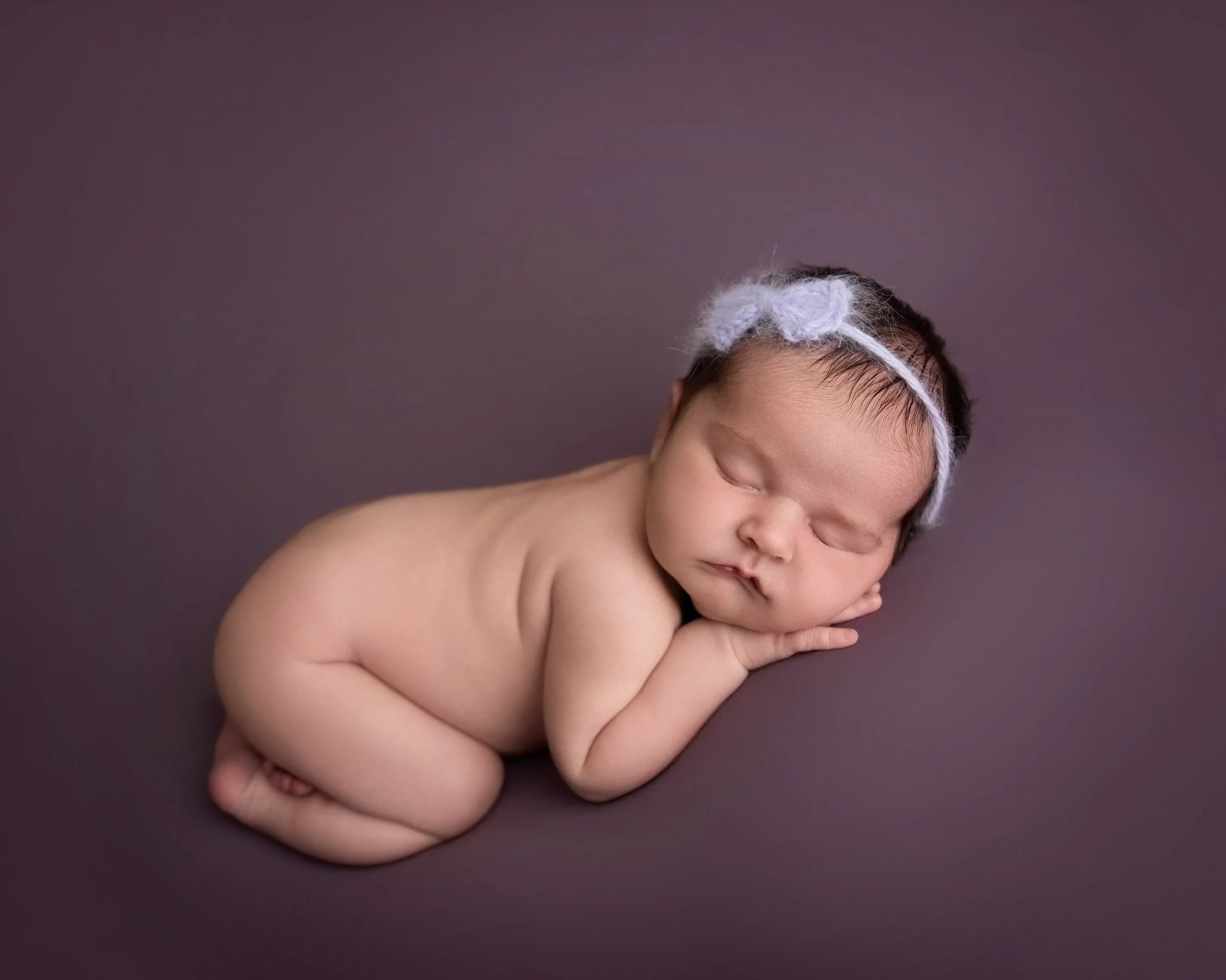 A sleeping newborn baby with a white headband, lying on a soft dark purple surface, curled up on their stomach with hands near face.