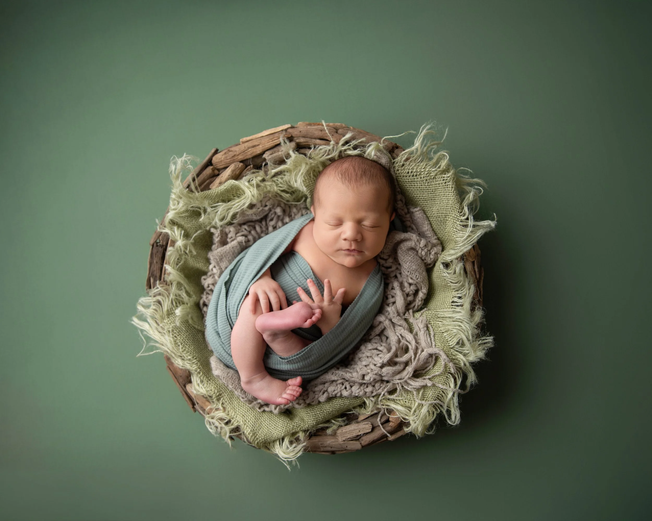Newborn baby peacefully sleeping, swaddled in a teal blanket, lying on soft layered fabrics in a woven basket on a green background.