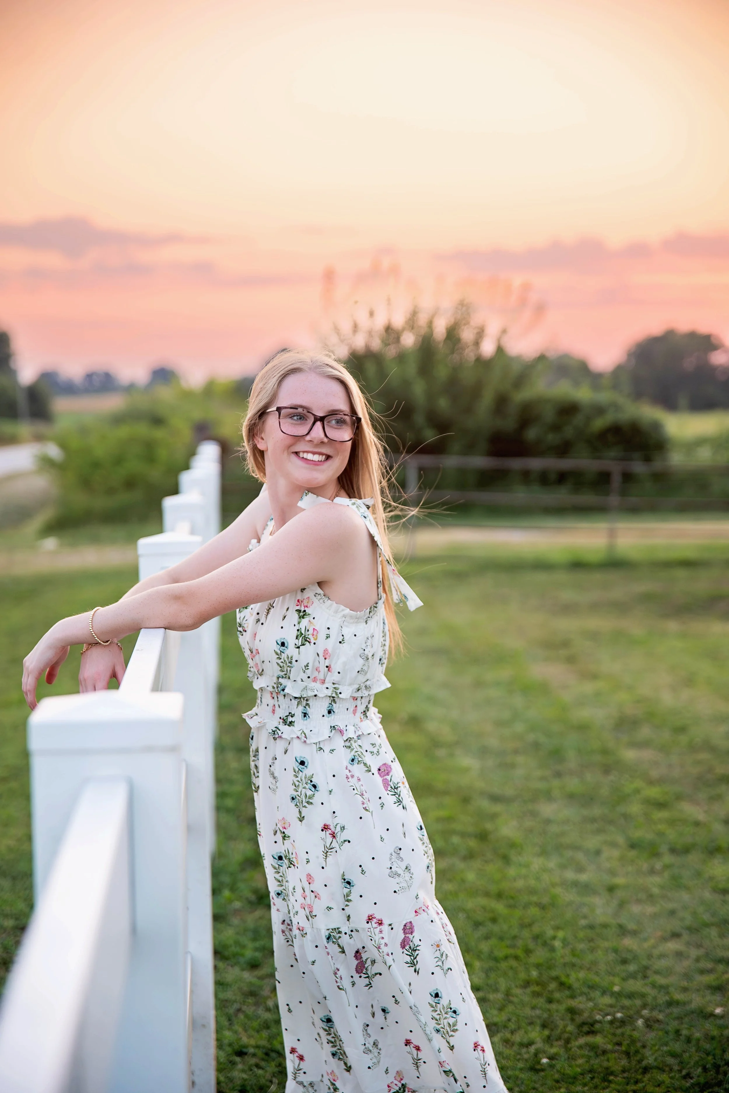 A young woman with blonde hair and glasses leaning on a white fence, smiling with a sunset sky in the background.