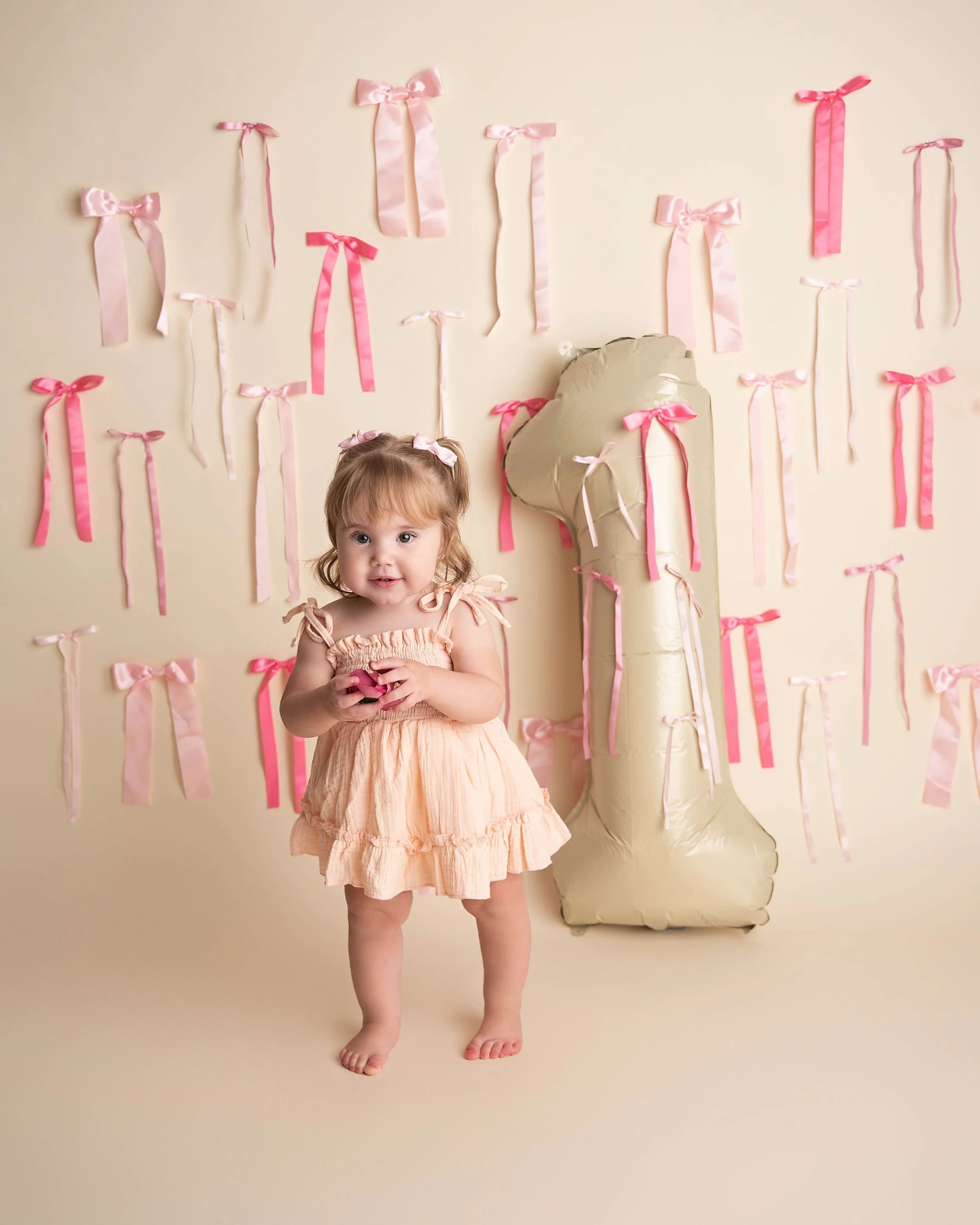 A young girl in a peach-colored dress standing in front of a decorated backdrop with pink ribbons and a large balloon shaped like the number 1.
