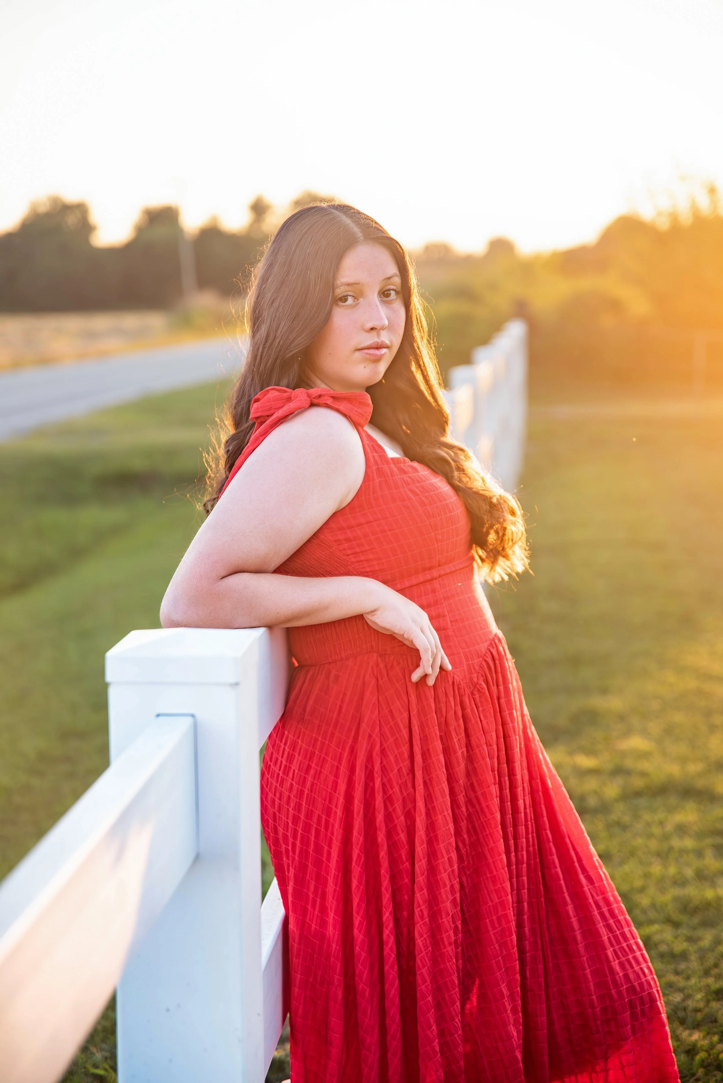 A young woman in a red dress leaning against a white fence outdoors during sunset, with green grass and trees in the background.