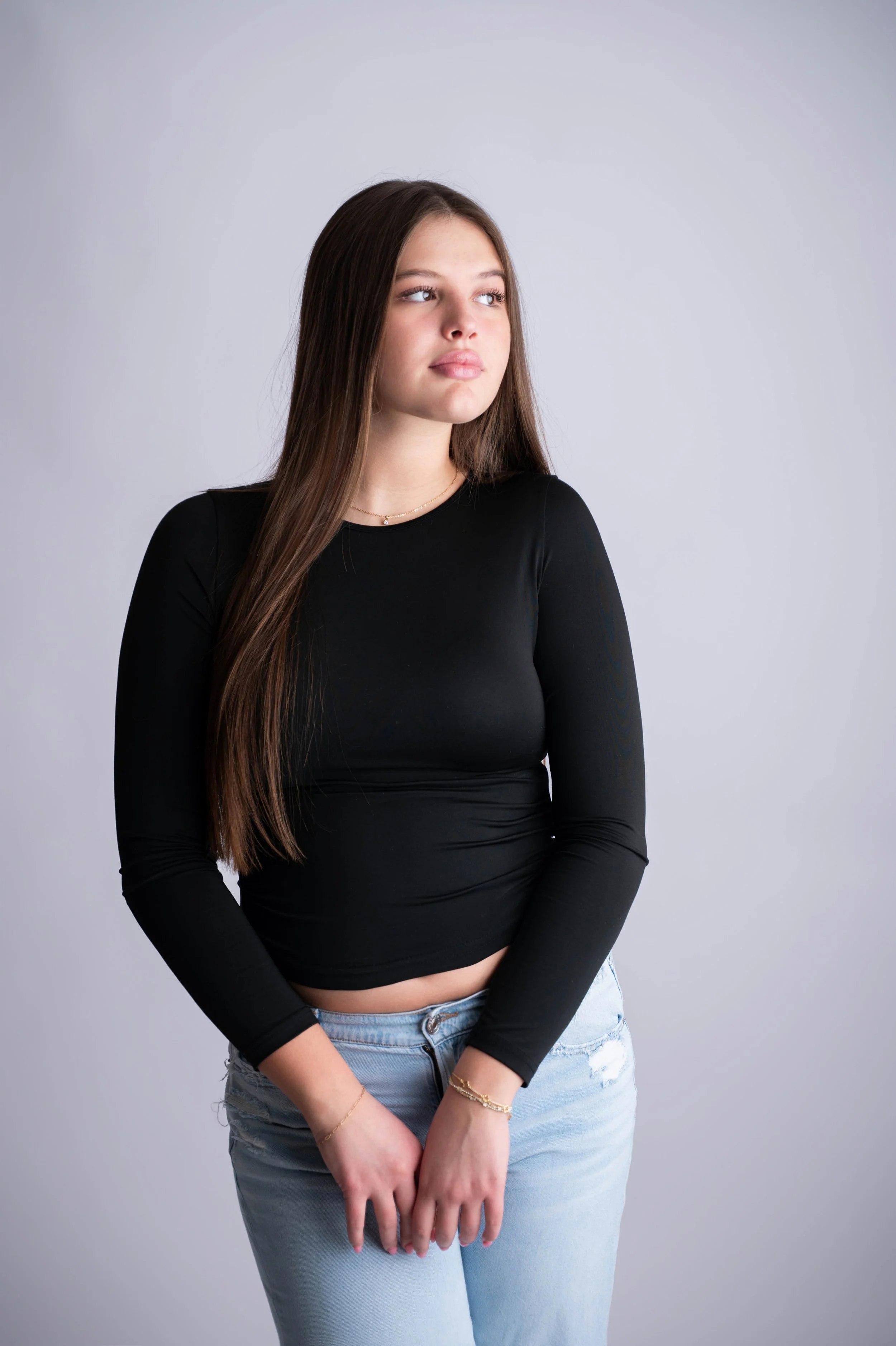 A young woman with long brown hair, wearing a black long-sleeve crop top and light blue jeans, standing against a plain gray background, looking slightly to the side.
