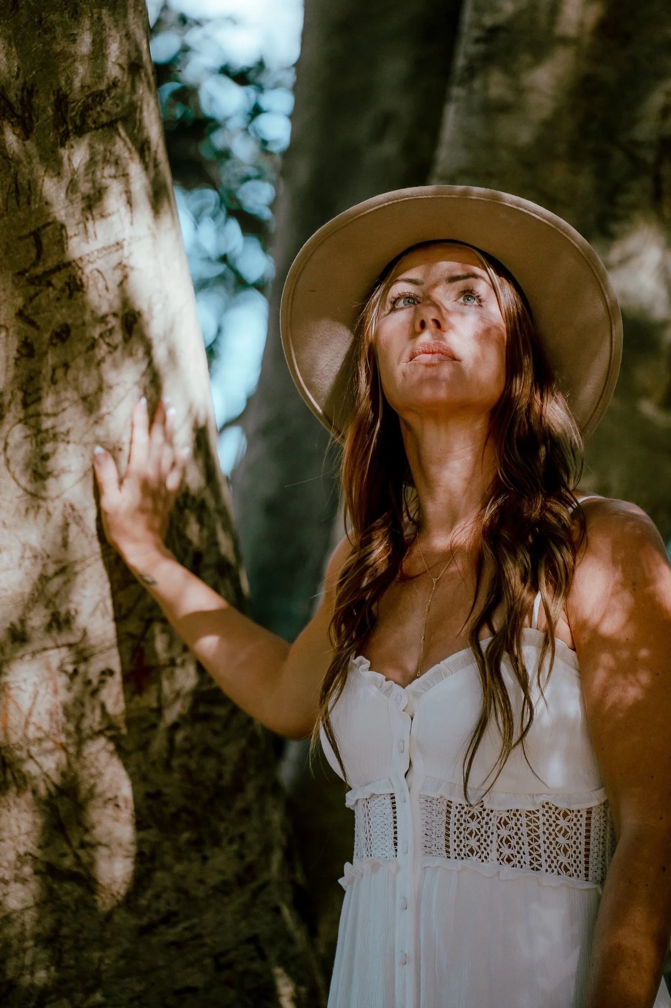 A woman with long wavy hair and a wide-brimmed hat is standing near a tree, looking upward, wearing a white dress with lace details.