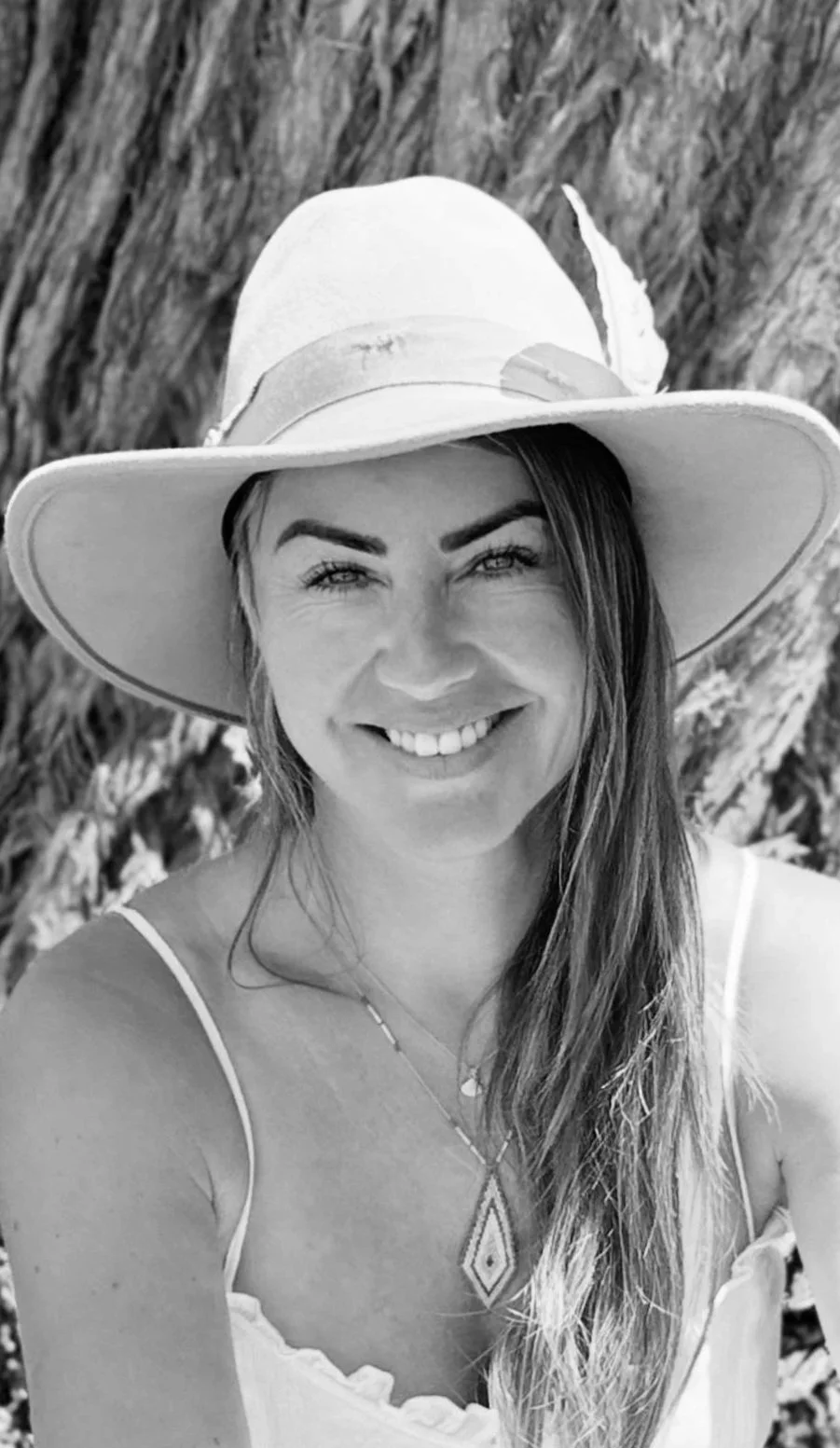 A woman smiling, wearing a large sunhat with a feather and a sleeveless top, outdoors with trees in the background.