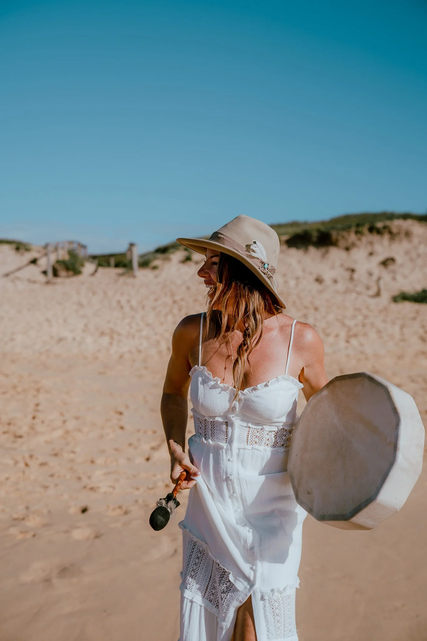Woman wearing a large sun hat and white dress, holding a round mirror and a small umbrella, standing on a sandy beach with dunes and a blue sky in the background.