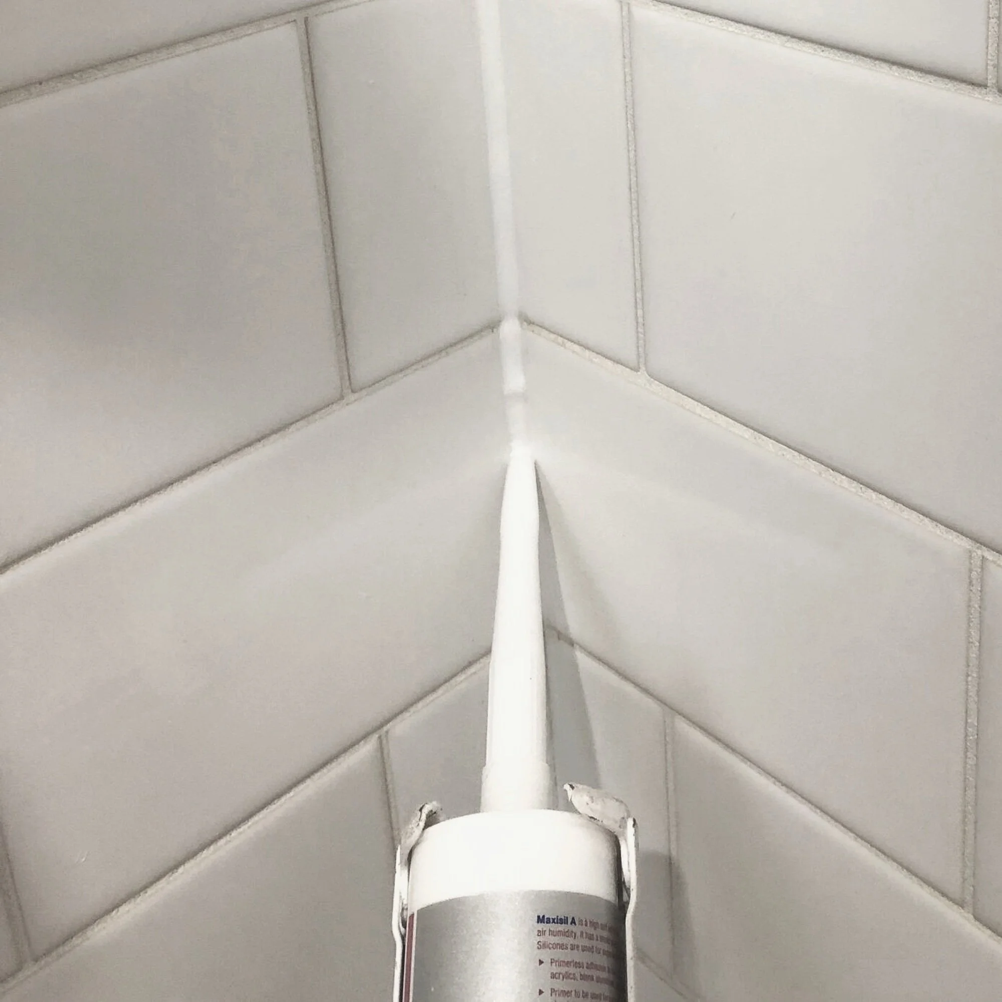 Close-up of a caulking gun applying white sealant to the corner of a tiled wall with white grout lines.