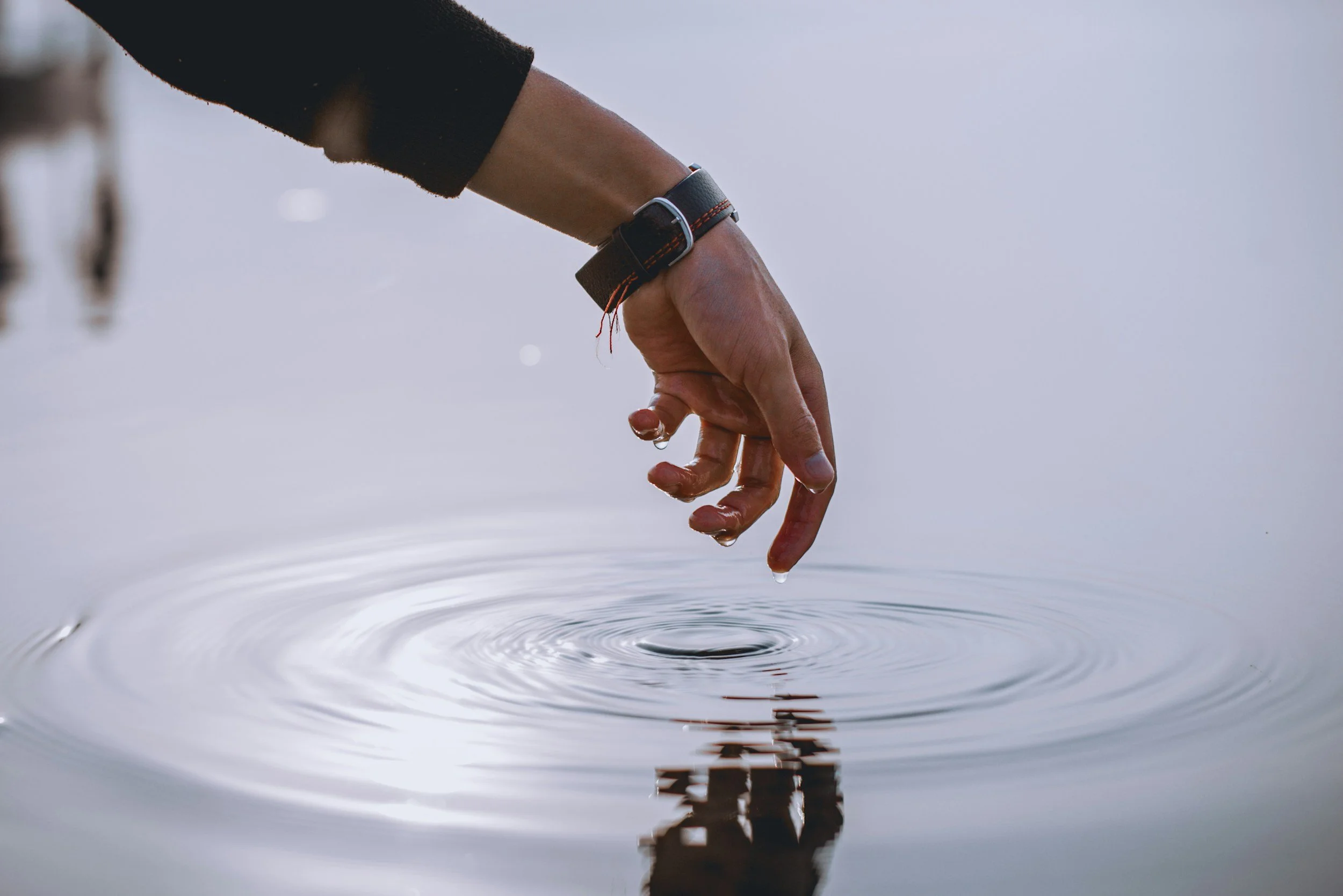 A person dips their fingers into calm water, creating ripples and droplets that hang from their fingertips.